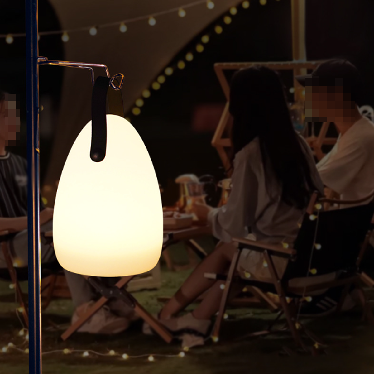 Eine leuchtend weiße Campinglaterne hängt. Menschen sitzen im Hintergrund unter Lichterketten an einem Tisch.