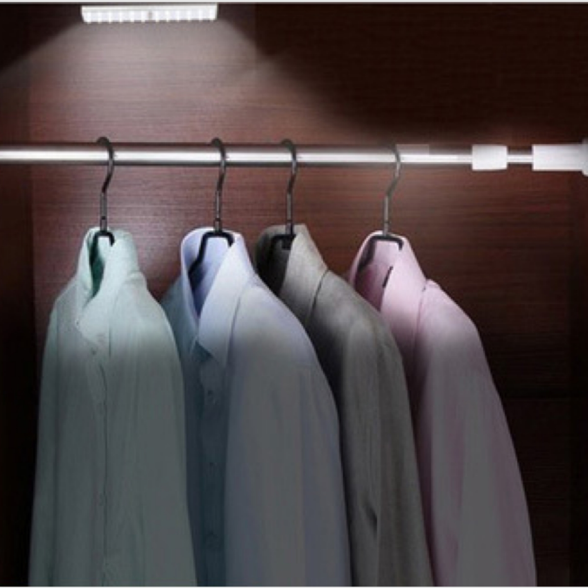 In einem Schrank hängen vier Hemden an Kleiderbügeln. Über den Hemden befindet sich eine Lampe.