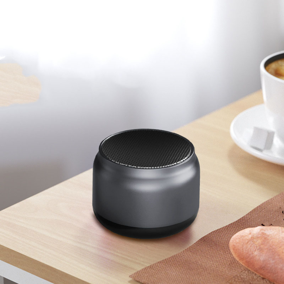 Ein kleiner, schwarzer, zylindrischer Lautsprecher auf einer Holzoberfläche mit Tasse und Gebäck.
