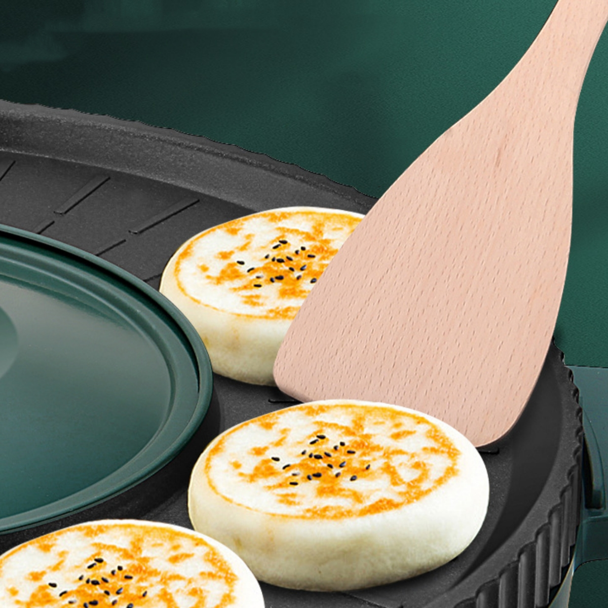 Pfannkuchen mit Sesam und Holzspatel auf einer Herdplatte, mit grünem Deckel im Hintergrund.
