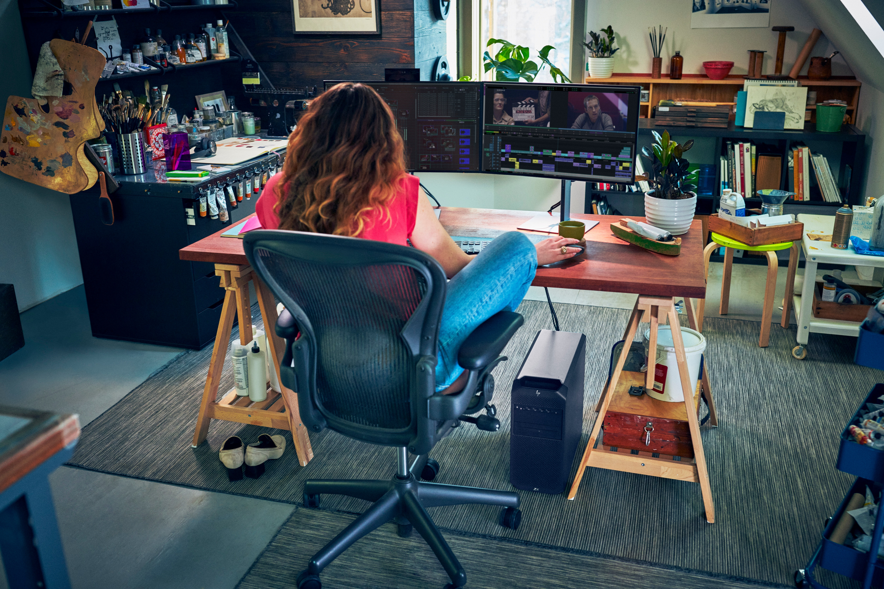 Eine Frau vor zwei Computerbildschirmen, die Videos bearbeitet. Schreibtisch mit Kunstutensilien, Computer und PC-Turm.
