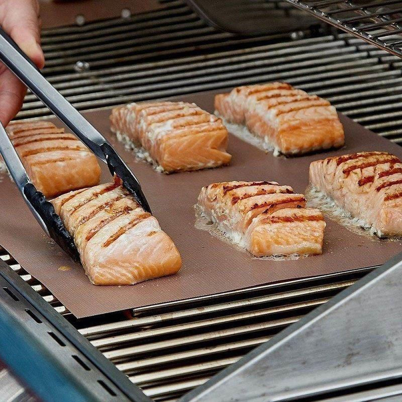 Lachsfilets auf einem Grill, werden mit Zangen gewendet. Grillspuren sichtbar. Braune Oberfläche.