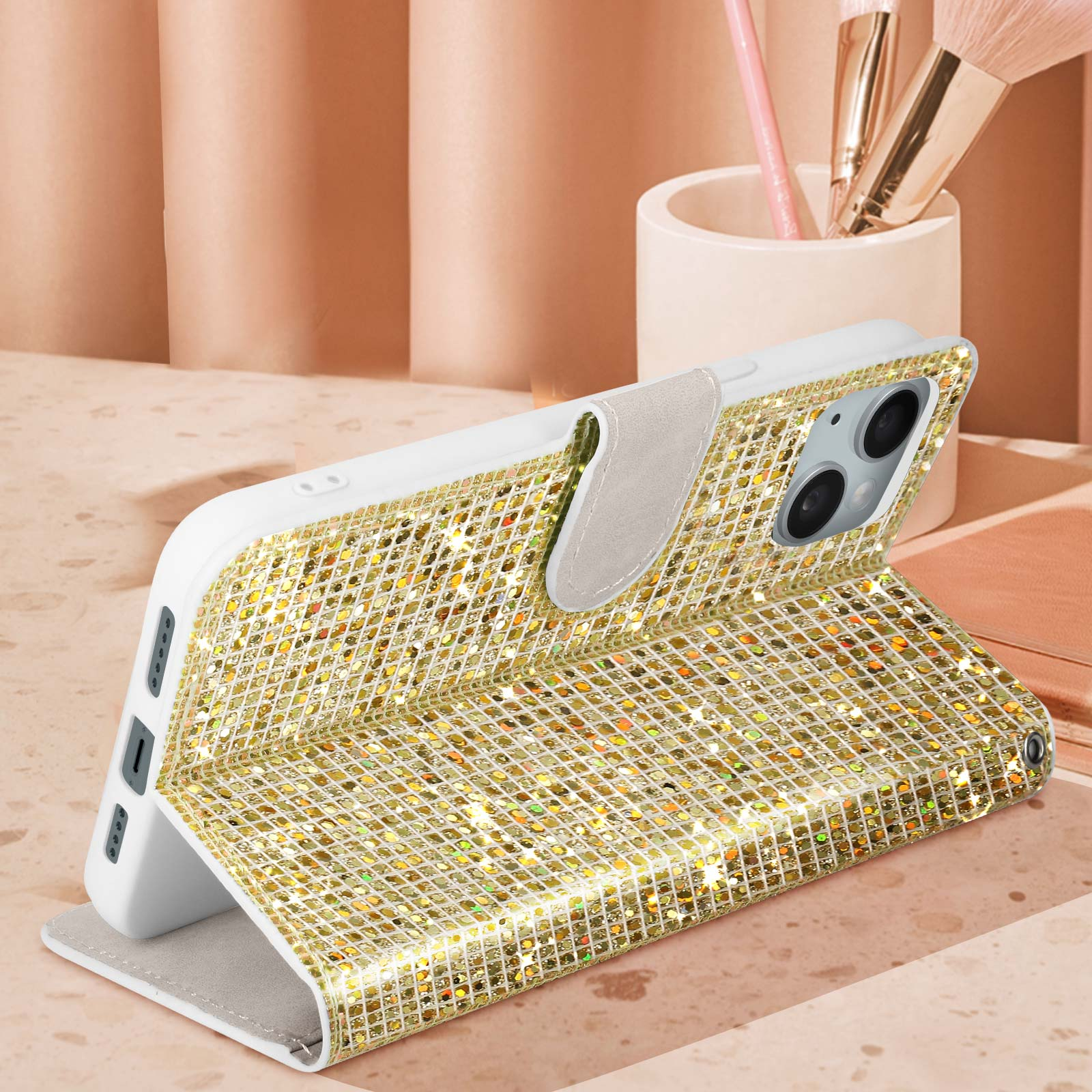 Offene goldene Handyhülle mit glitzernden Details. Hintergrund enthält Make-up-Pinsel und eine hellfarbige Oberfläche.