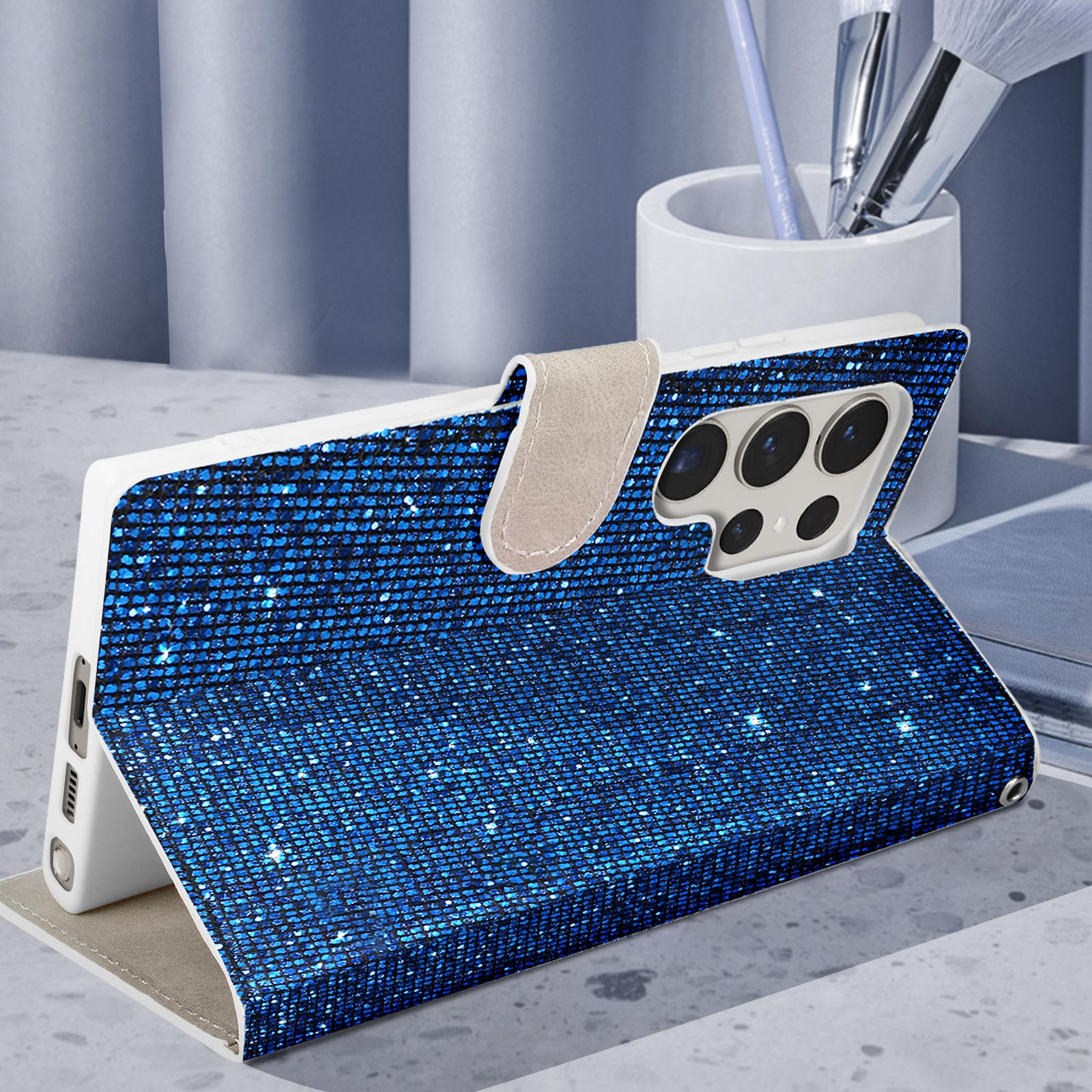 Custodia per telefono blu glitterata, aperta e inclinata. Telefono bianco all'interno. Supporto, pennelli sullo sfondo.