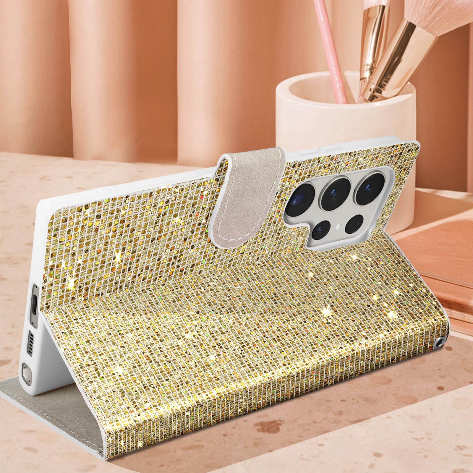Custodia per telefono glitterata oro, aperta, che mostra il telefono. La custodia è su una superficie rosa con pennelli per il trucco.