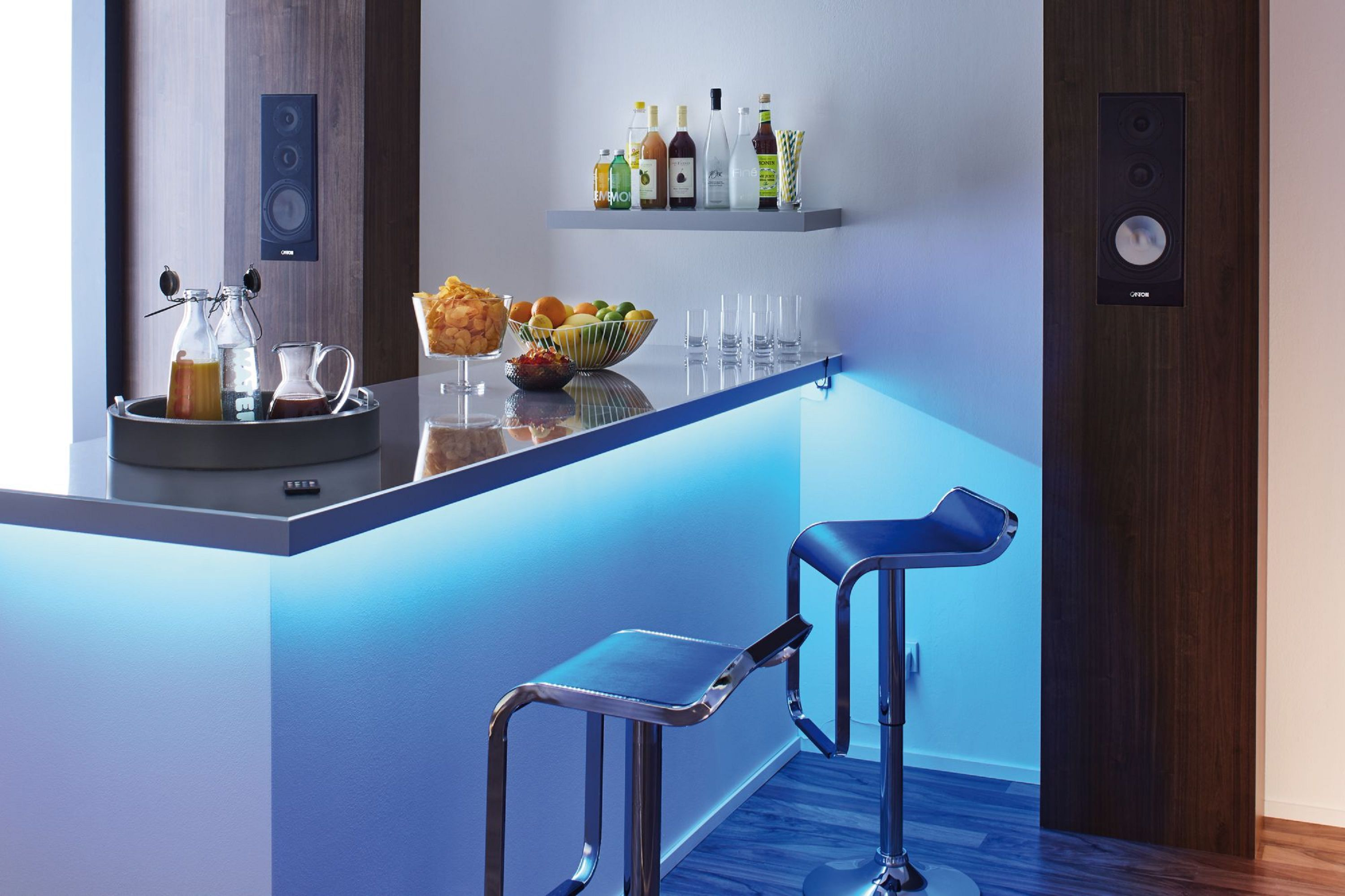 Moderne Bar mit blauer Beleuchtung, Hockern und Lautsprechern. Verschiedene Flaschen und Snacks auf Regalen.