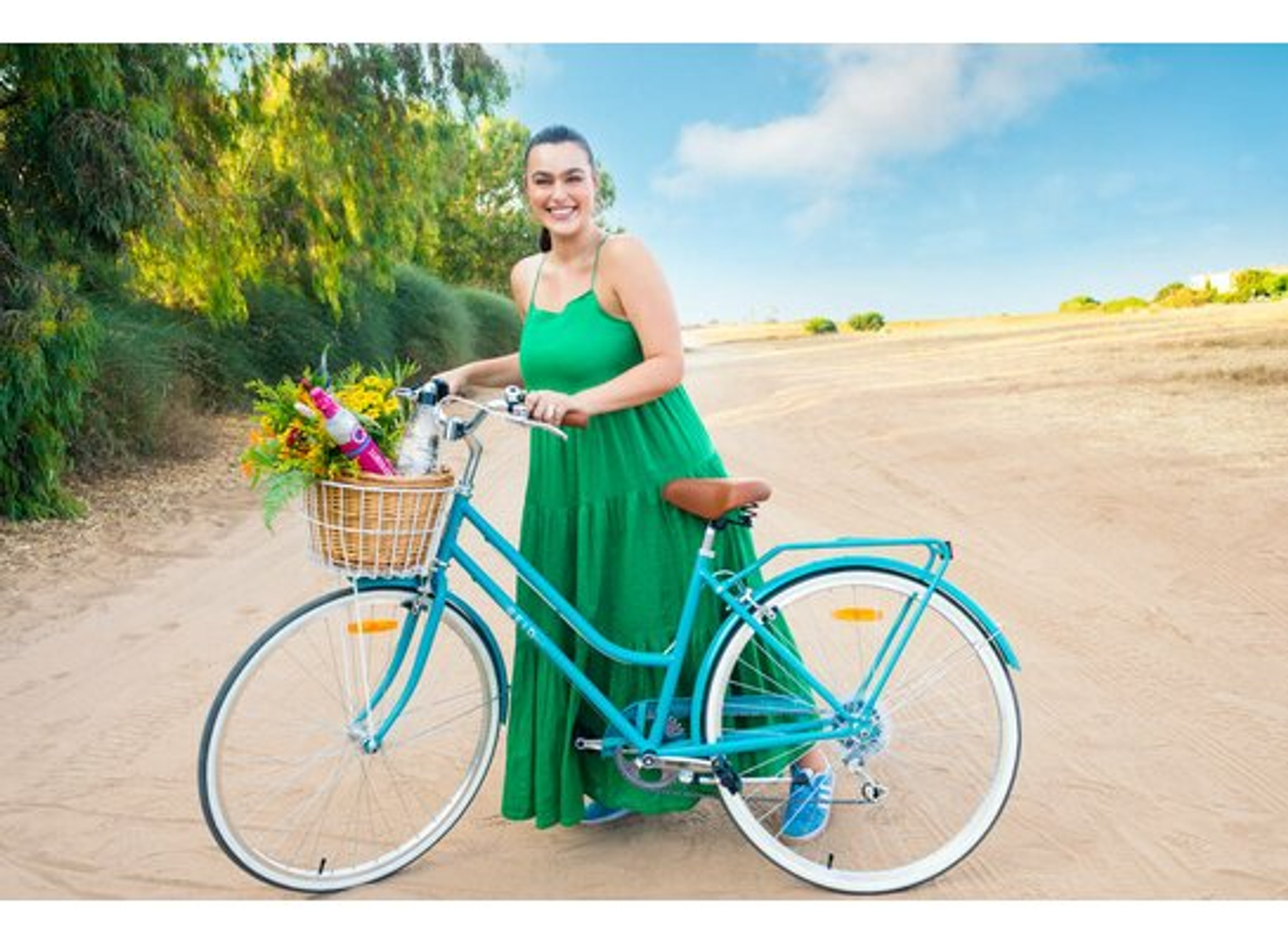 Donna con bici, vestito verde, cesto di fiori, cielo blu, strada.