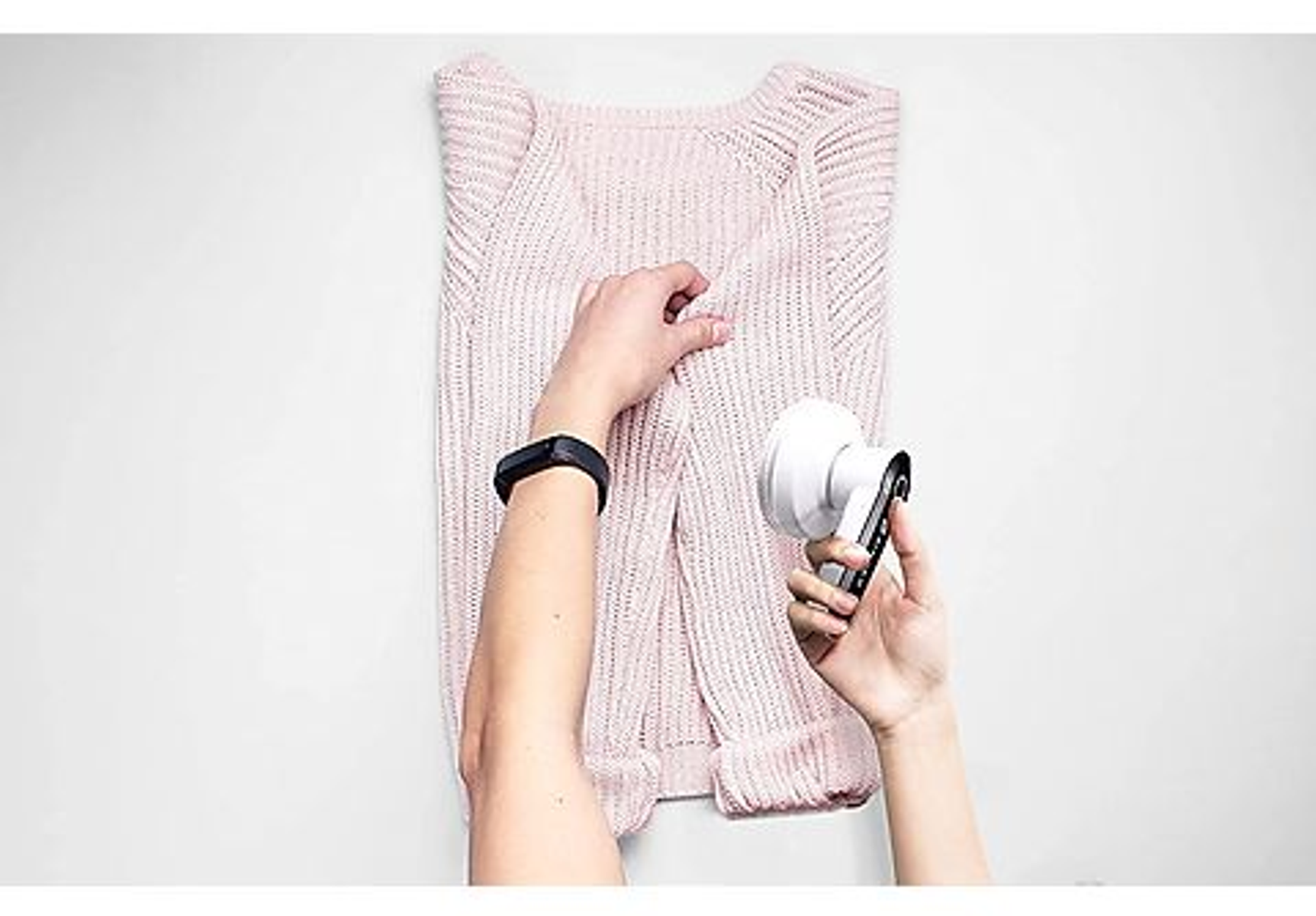 Hände, die eine Fusselentfernungsmaschine auf einem rosa Pullover verwenden. Der Pullover liegt flach auf einer weißen Oberfläche.