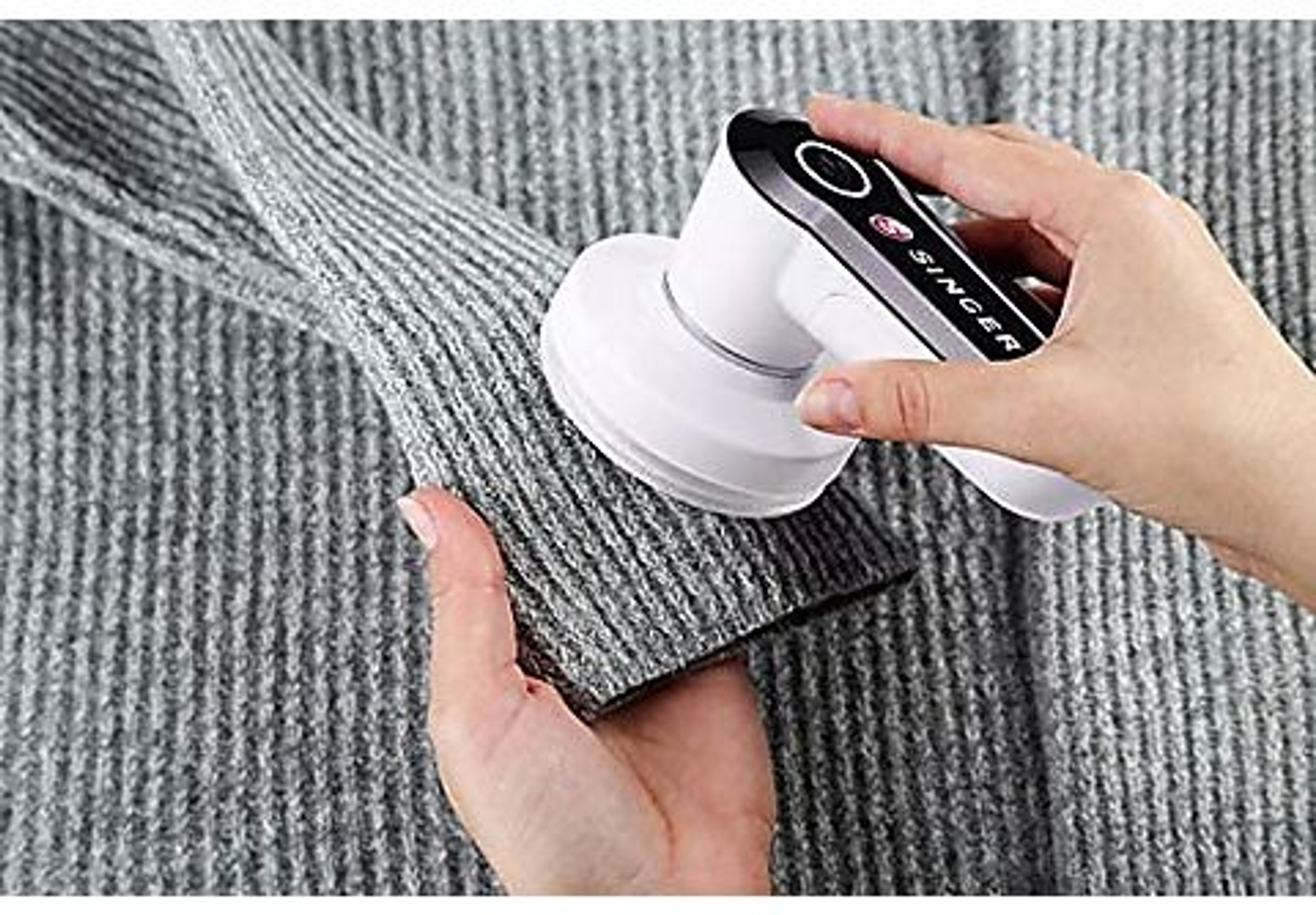 Eine Person, die einen Stoffrasierer an einem grauen Pullover verwendet. Der Rasierer ist weiß und schwarz.