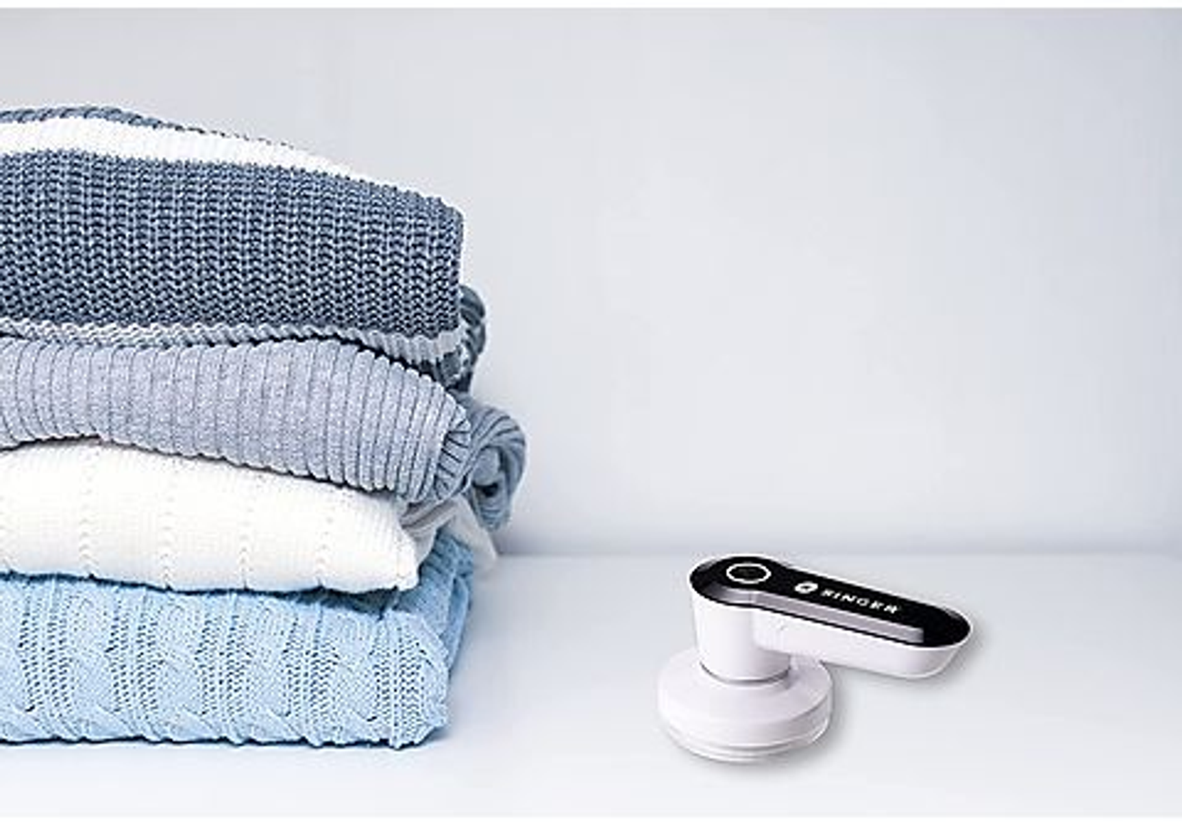 Stapel gefalteter blauer, grauer und weißer Pullover mit einem Fusselentferner daneben.