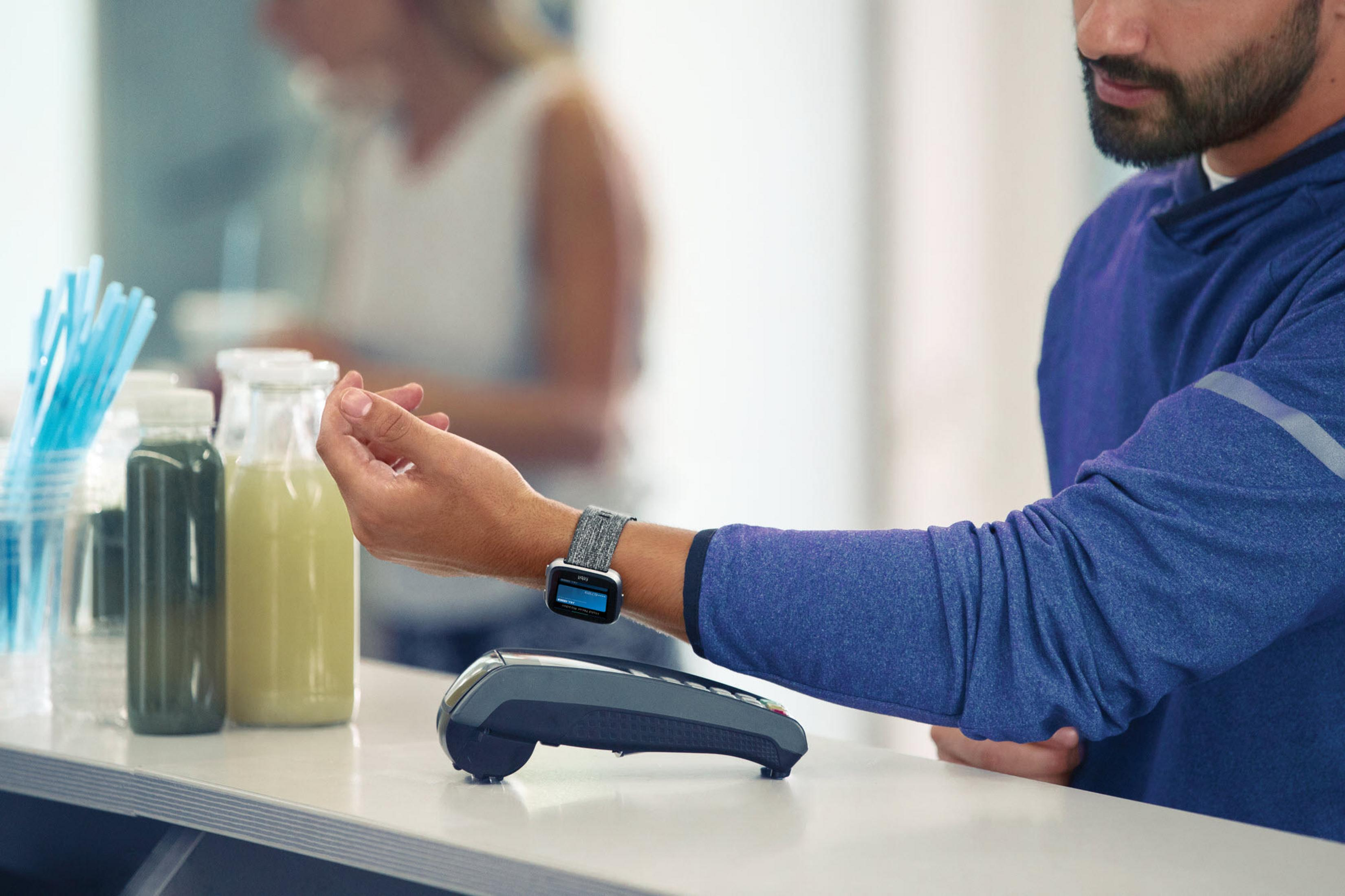Eine Person zahlt mit einer Smartwatch an einem Tresen. Ein Zahlungsgerät und Getränke sind ebenfalls sichtbar.