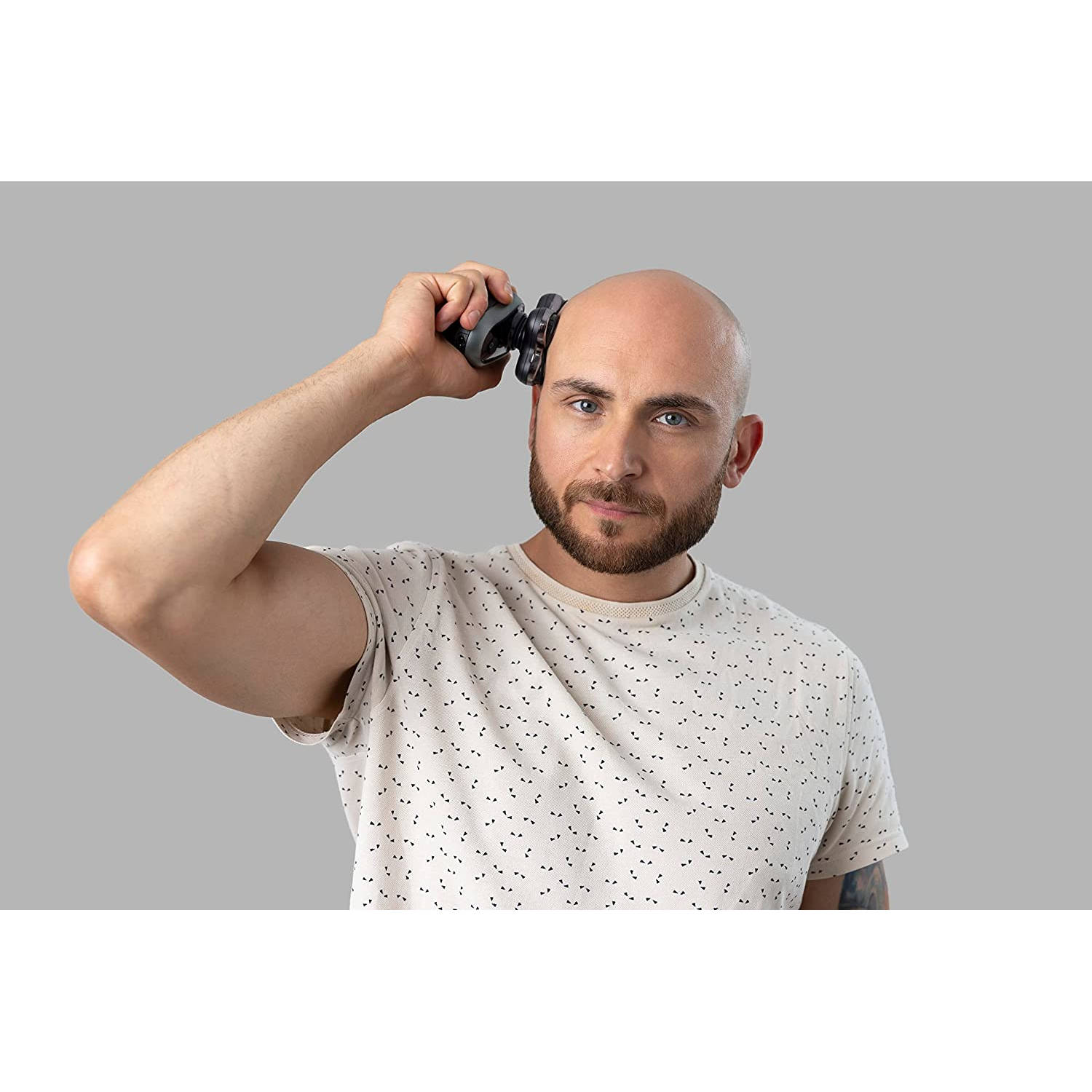 Ein Mann rasiert sich mit einem schwarzen Elektrorasierer den Kopf. Er trägt ein T-Shirt und schaut den Betrachter an.