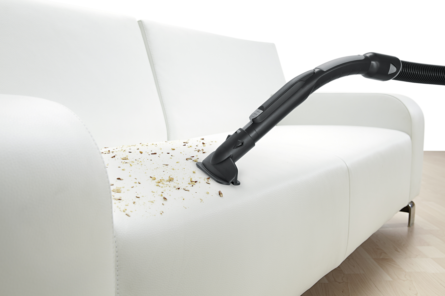 Eine Staubsaugerdüse reinigt ein weißes Sofa. Auf dem Kissen sind Ablagerungen verstreut.