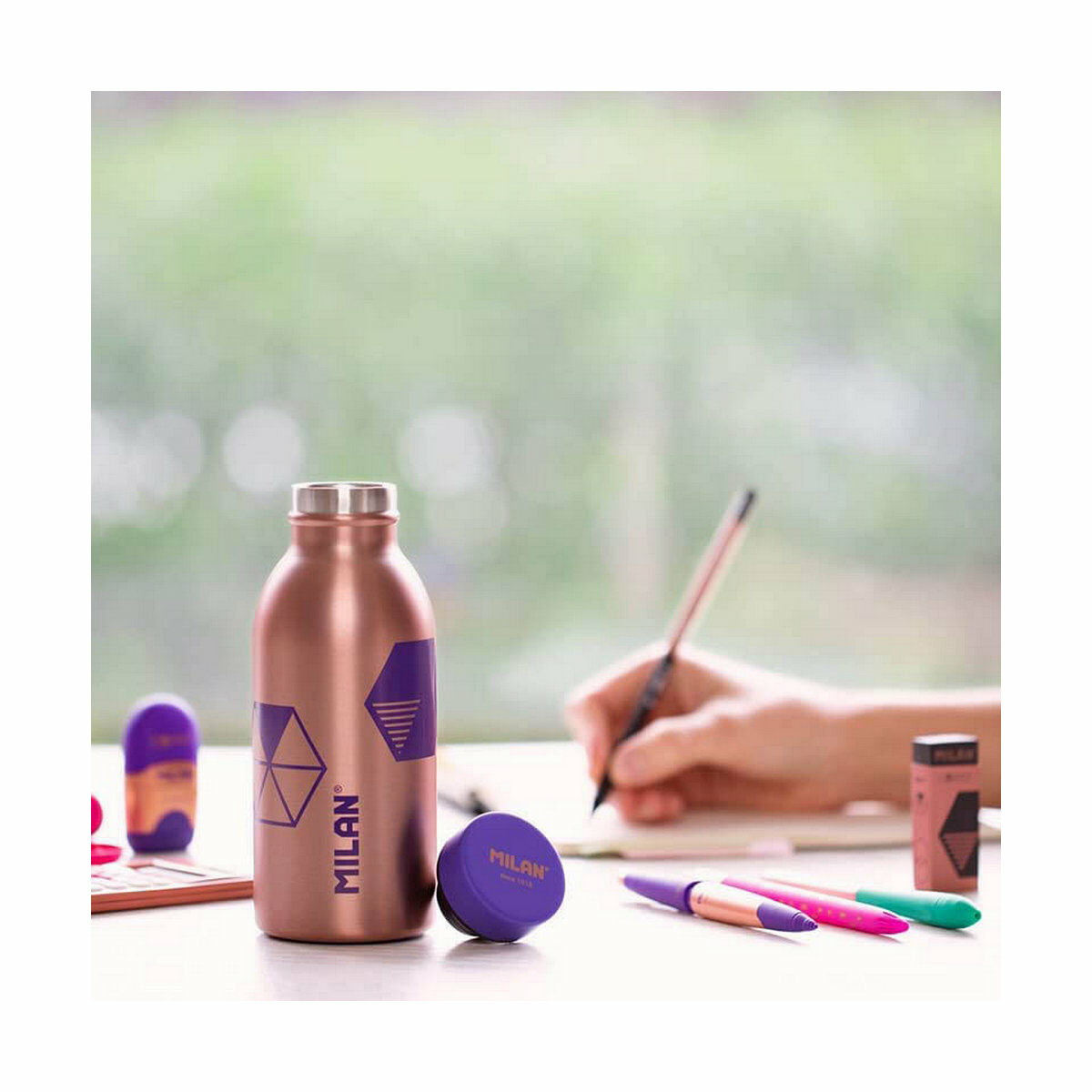 Eine kupferfarbene Wasserflasche, Stifte und ein Radiergummi liegen auf einem Tisch. Eine Hand schreibt mit einem Stift. Im Hintergrund ist ein Fenster zu sehen.
