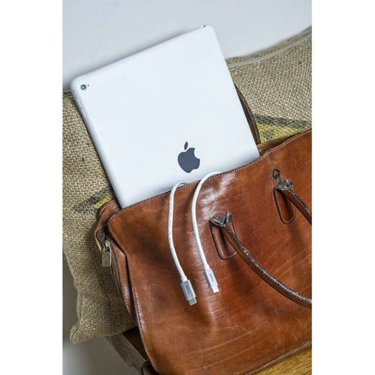 Ein weißes iPad und ein Ladekabel in einer braunen Ledertasche, auf einem Jutekissen.