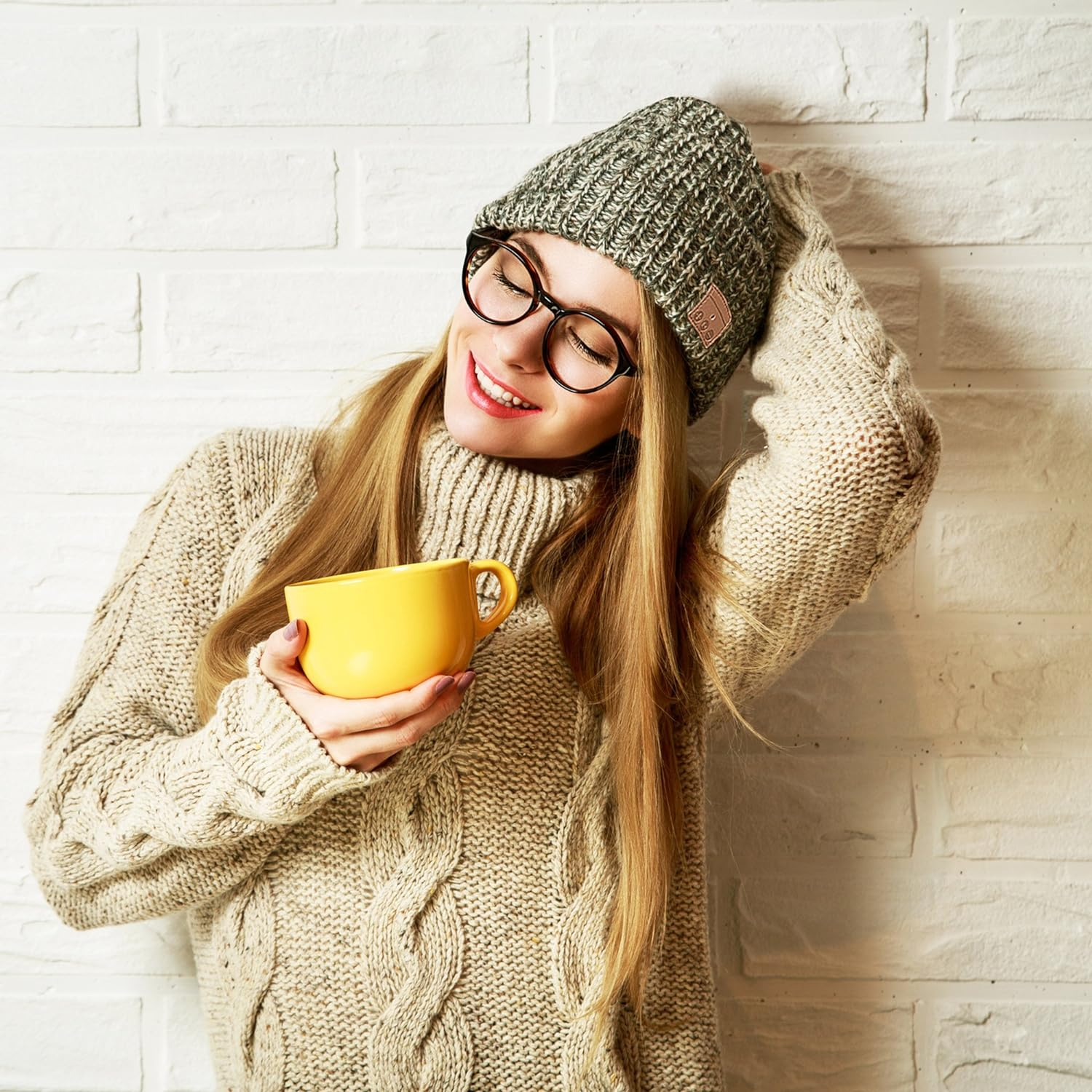 Frau mit Brille und Mütze, hält eine gelbe Tasse, lächelt, trägt einen Pullover.