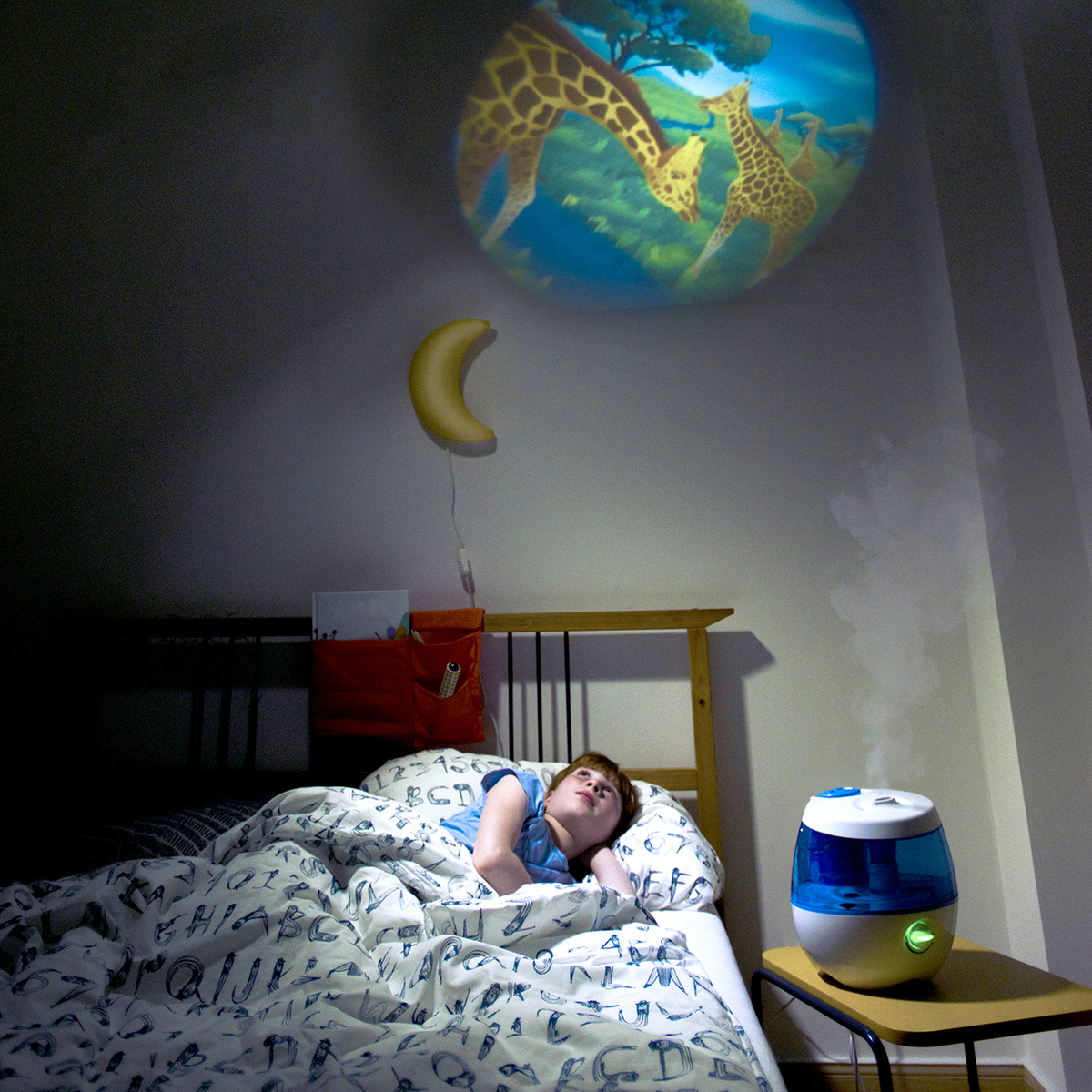 Ein Junge liegt im Bett und schaut auf eine Giraffenbildprojektion an der Decke. Neben dem Bett steht ein Luftbefeuchter.