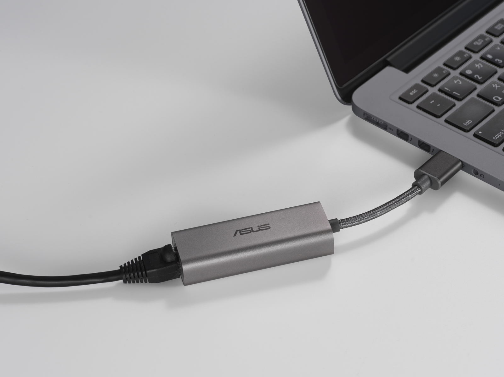 Ein grauer ASUS-Adapter, der mit einem Laptop und einem schwarzen Kabel auf weißer Oberfläche verbunden ist.