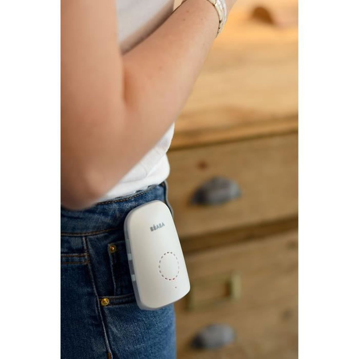 Ein weißer Baby-Monitor, der an der Jeans einer Frau befestigt ist, im Hintergrund eine Holzkommode.