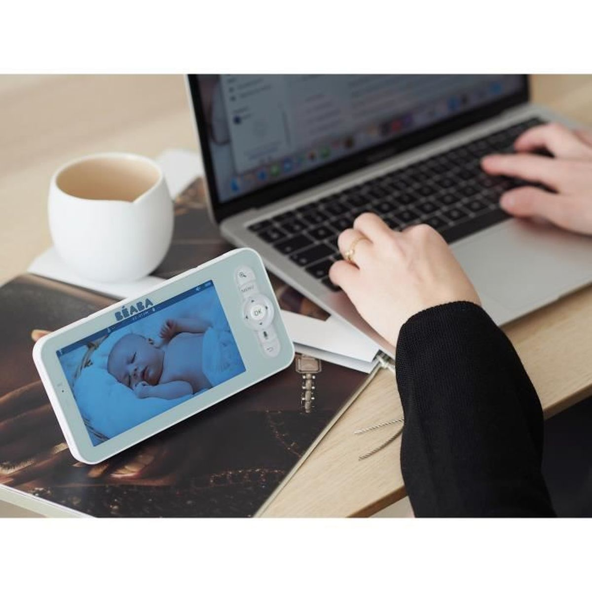 Un baby monitor con un bambino che dorme, accanto a un laptop su una scrivania. Una persona sta usando il laptop.