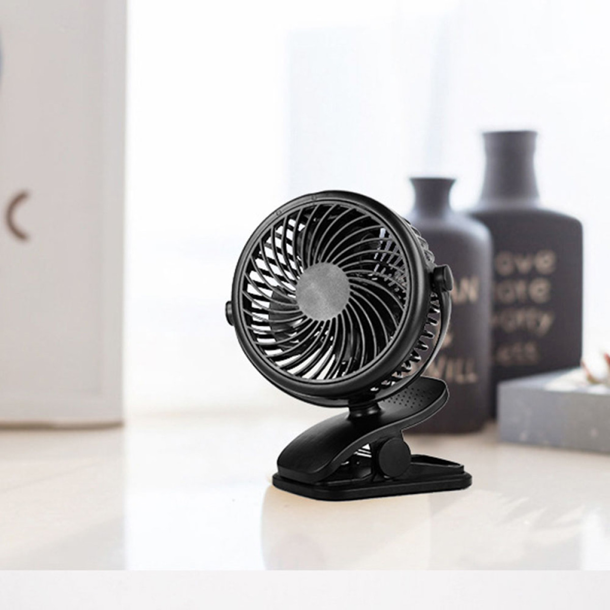 Ein kleiner schwarzer Ventilator mit Clip auf einer hellen Oberfläche. Zwei schwarze Vasen im Hintergrund.