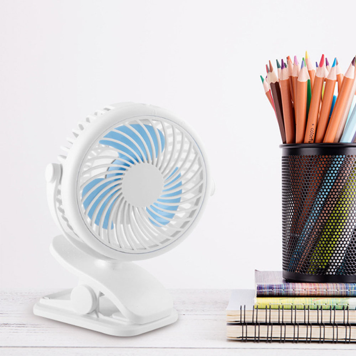 Weißer Ventilator mit blauen Flügeln auf einem Schreibtisch mit Stiften und Notizbüchern.