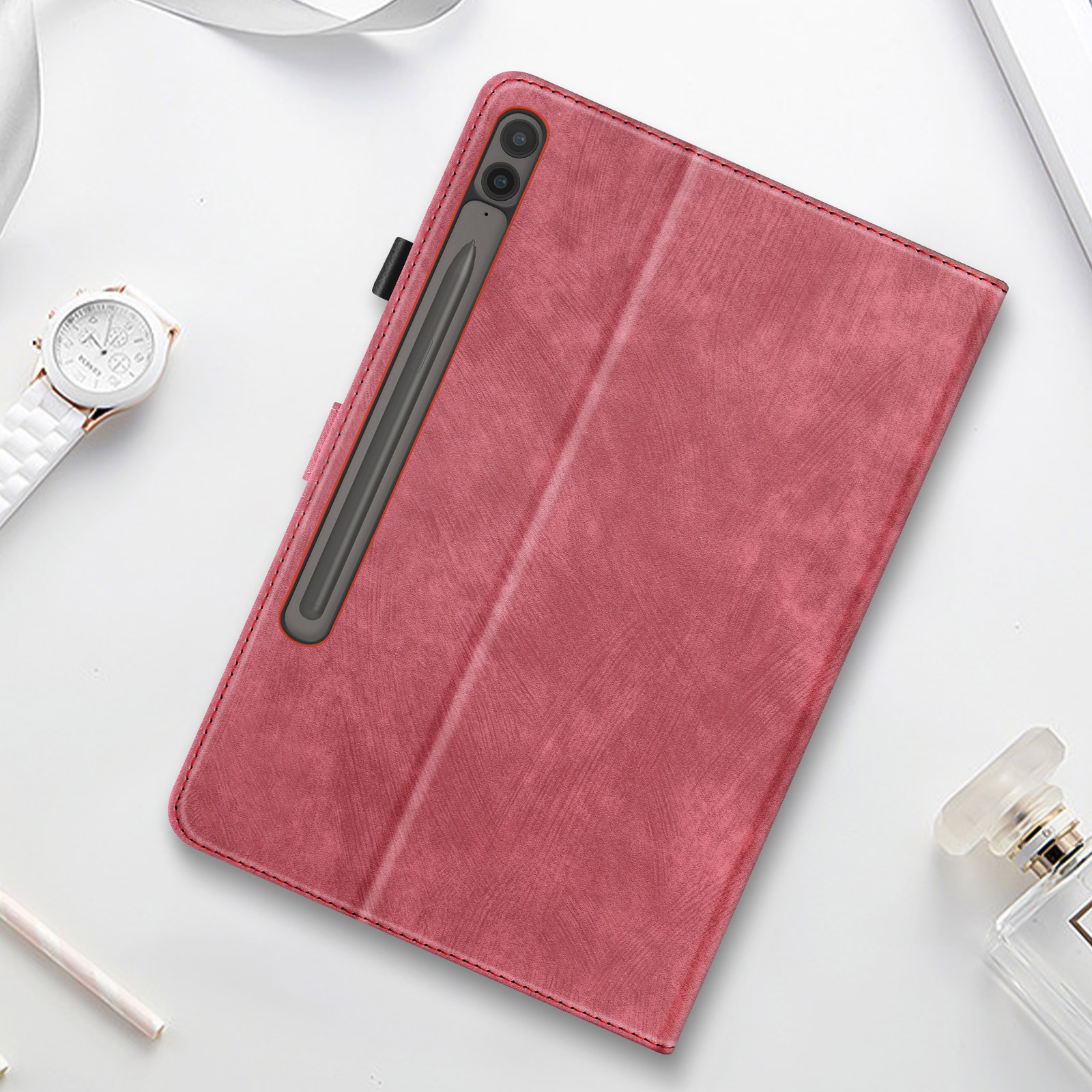 Custodia per tablet rosa con stilo, orologio, profumo e penna su superficie bianca.