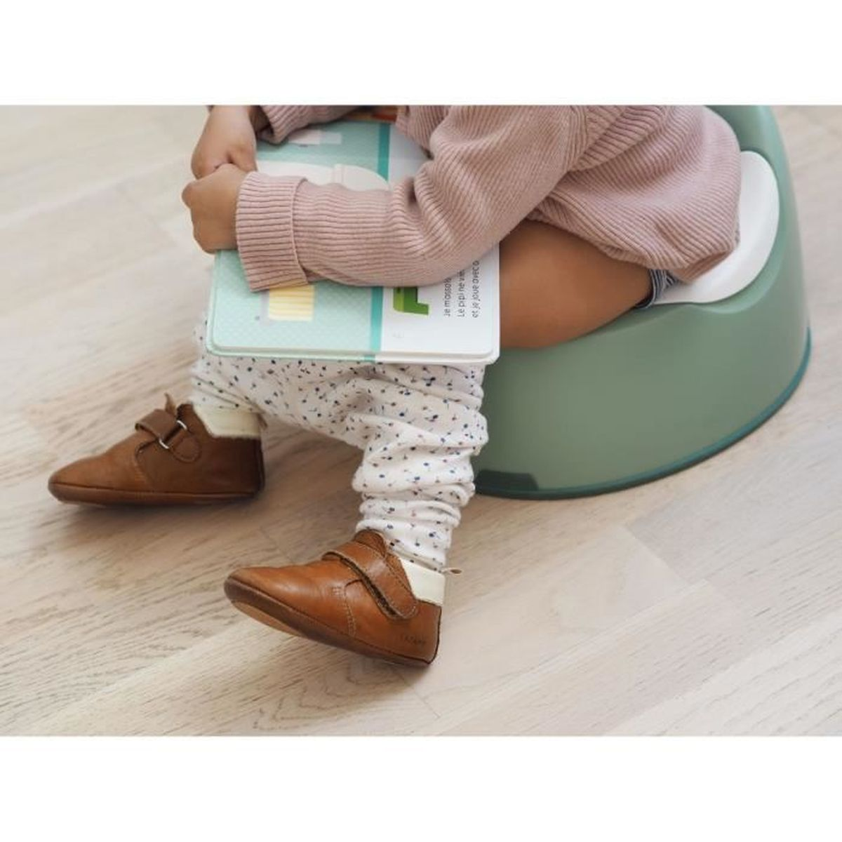 Ein Kind sitzt auf einem grünen Töpfchen und liest ein Buch, trägt Schuhe.