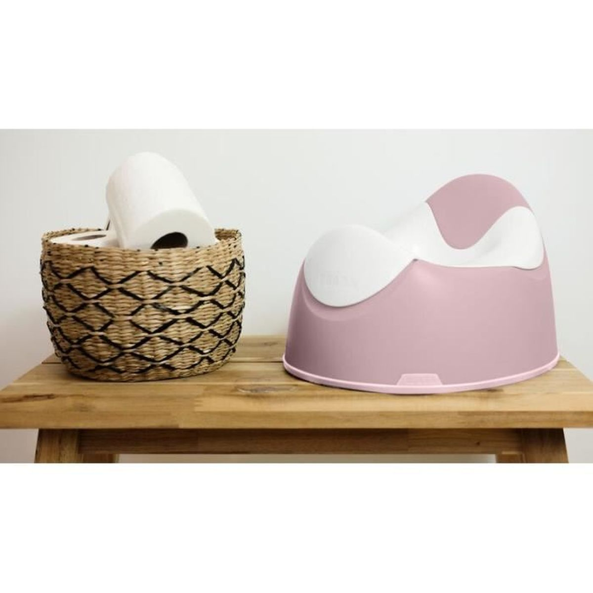 Rosa-weißes Töpfchen mit einer Rolle Toilettenpapier in einem Weidenkorb. Die Anordnung ist auf einer Holzoberfläche.