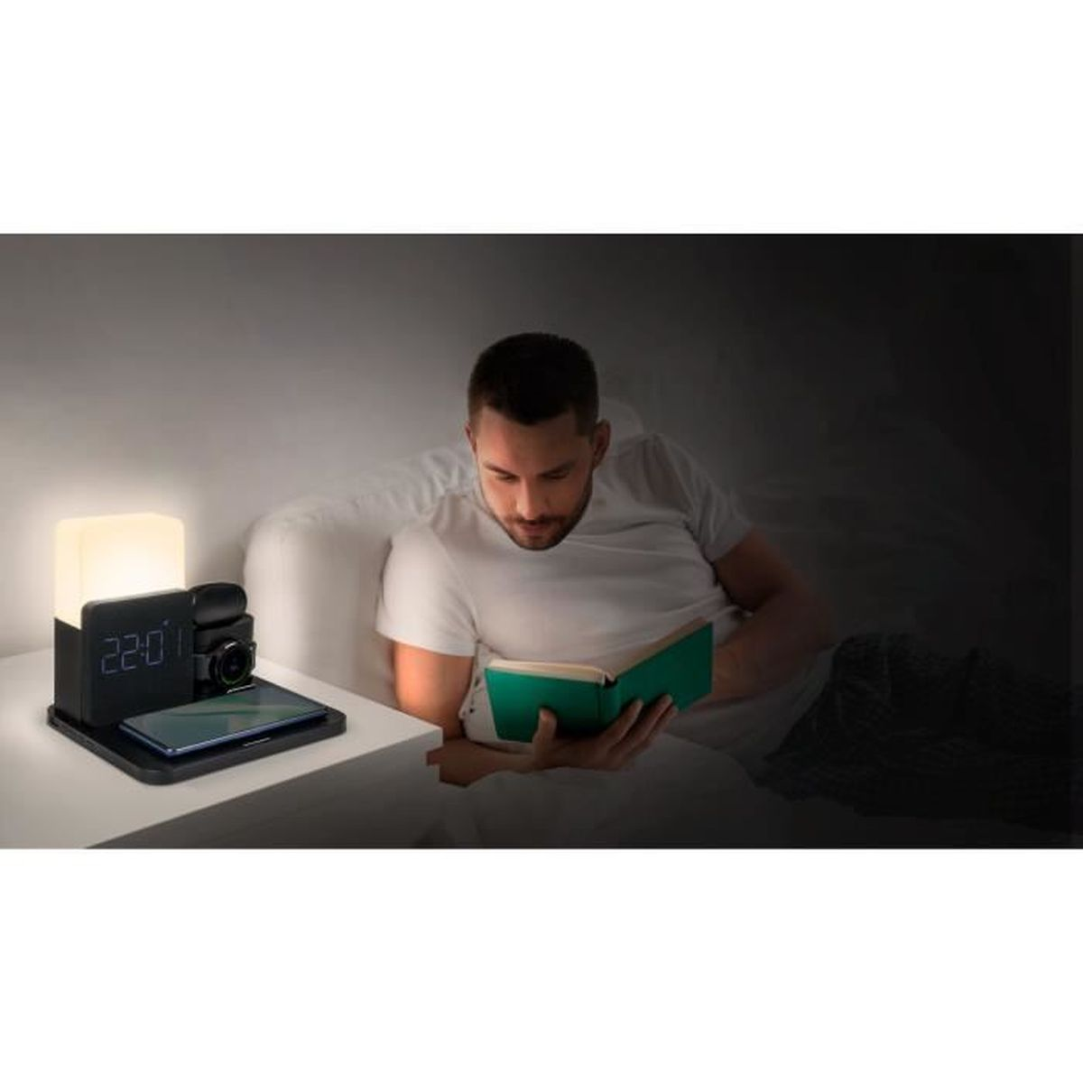 Mężczyzna czytający książkę w łóżku, ze stacją ładującą i lampą. Ciemne tło.