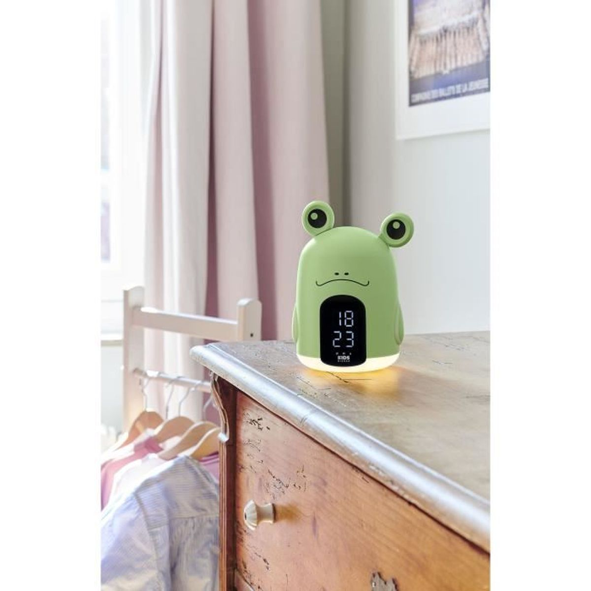 Luz nocturna/despertador con forma de rana verde sobre una superficie de madera, cortina rosa detrás.