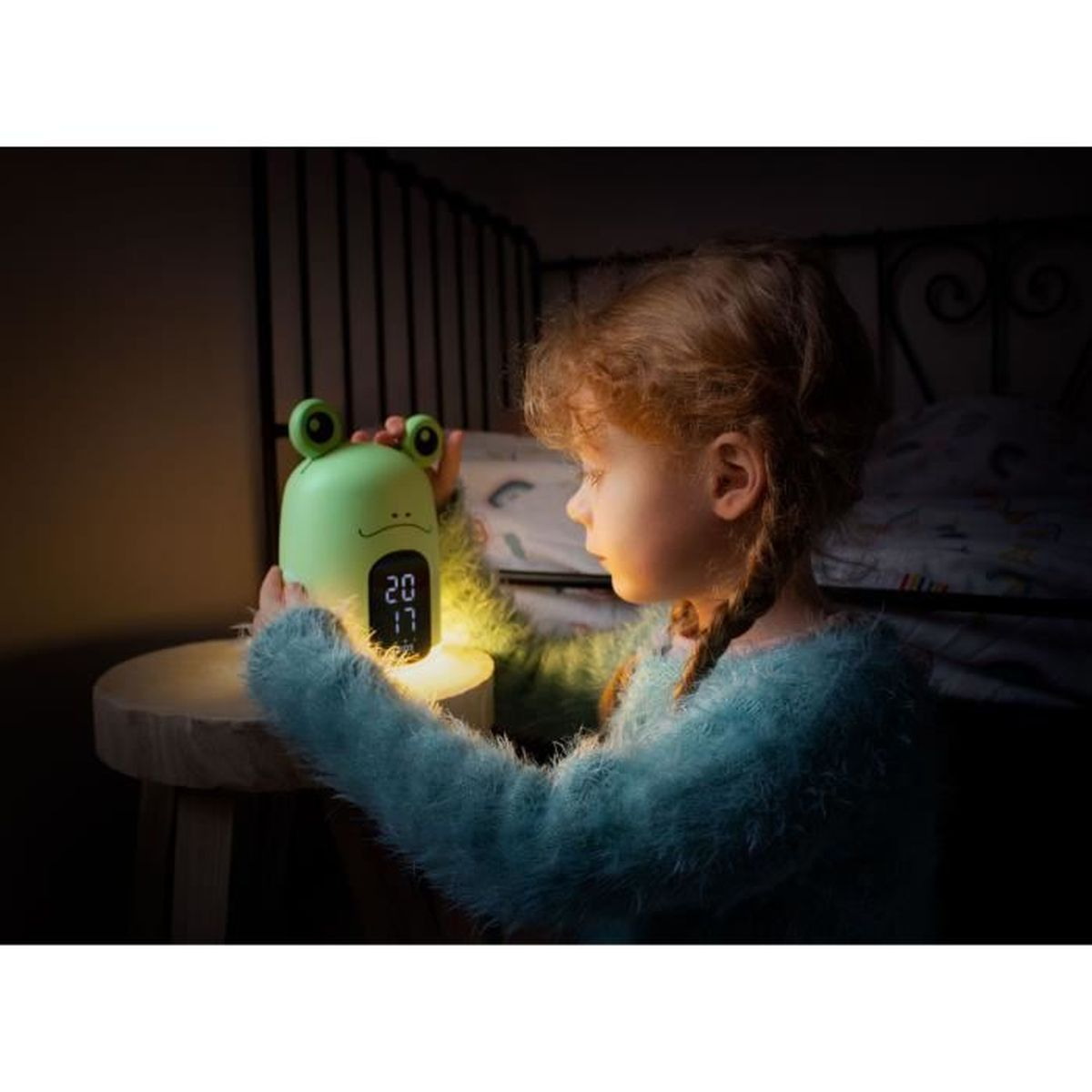 Una niña mirando una luz de noche verde con forma de rana. El reloj muestra 20:17.