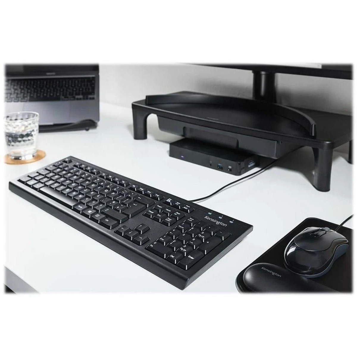 Czarny zestaw komputerowy: klawiatura, mysz, stojak na monitor z urządzeniami, białe biurko.