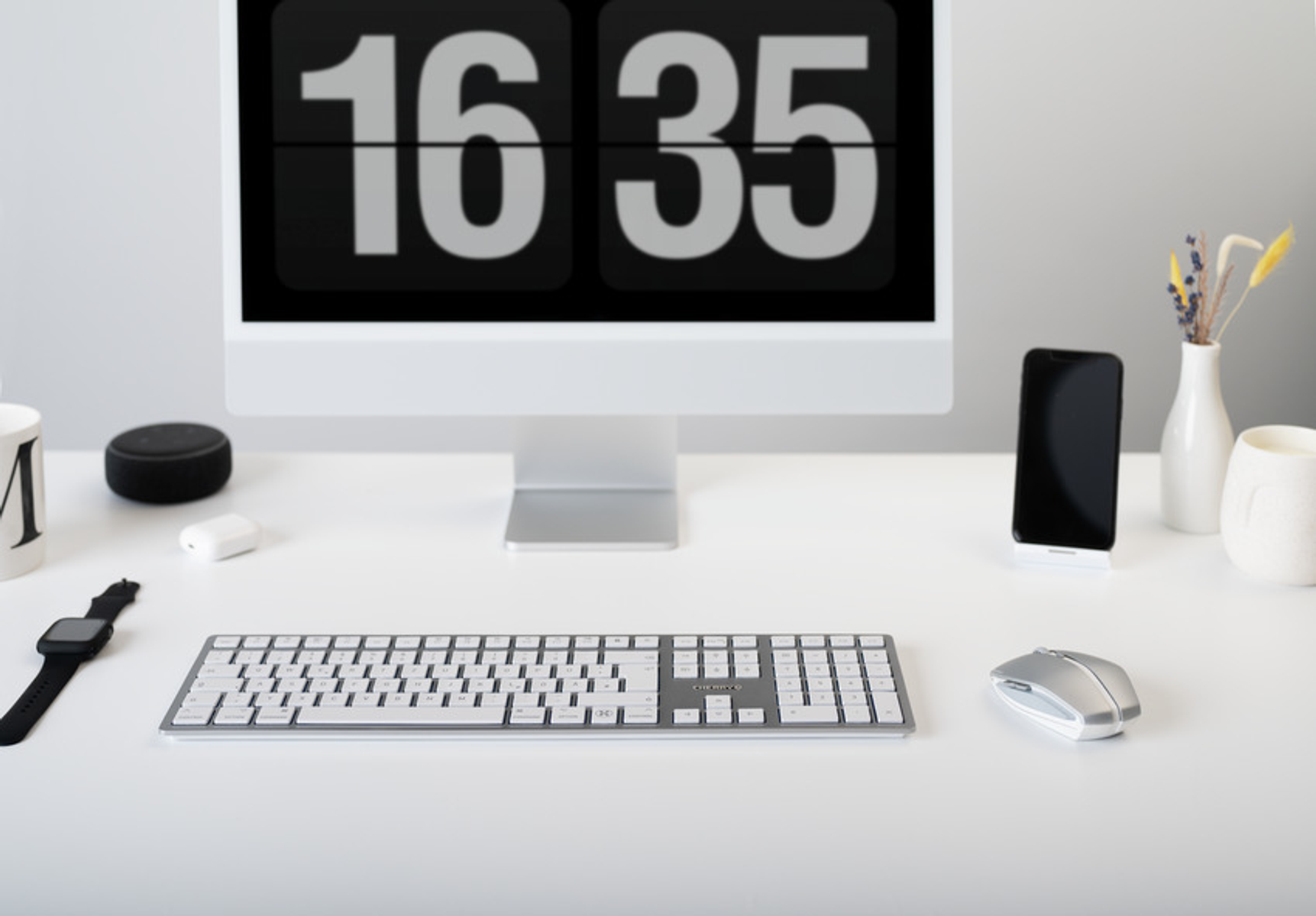 Wit bureau met computer, klok, telefoon en andere accessoires. De klok geeft 16:35 aan.