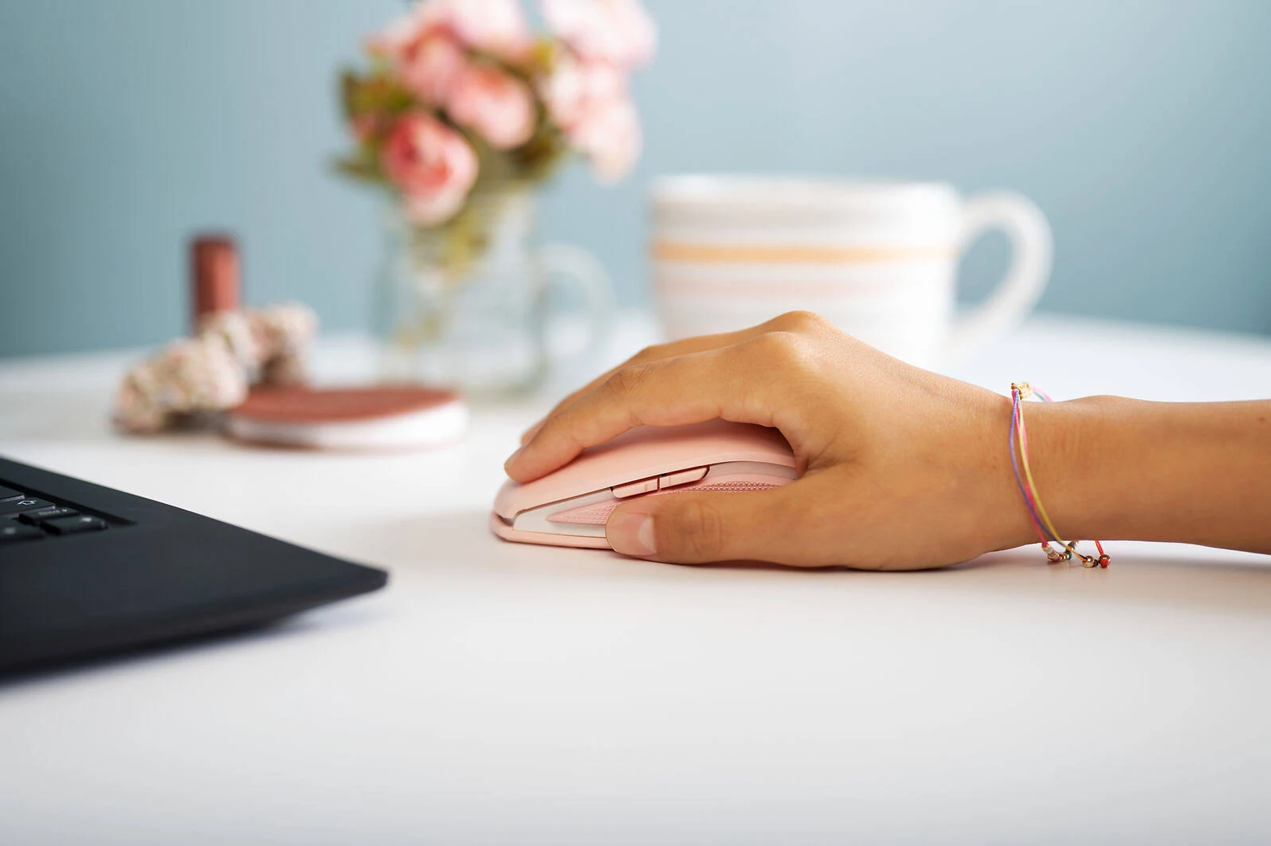 A hand using a pink wireless mouse next to a laptop on a white desk.