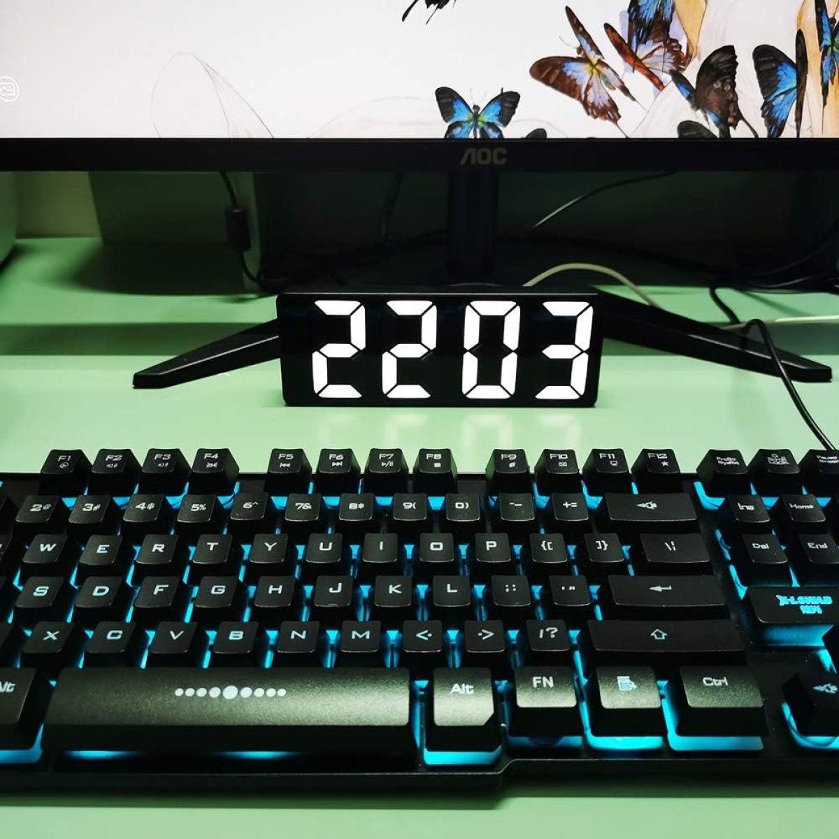 Tastatur mit blauer Hintergrundbeleuchtung, Digitaluhr mit 22:03 und Monitor mit Schmetterlingen.