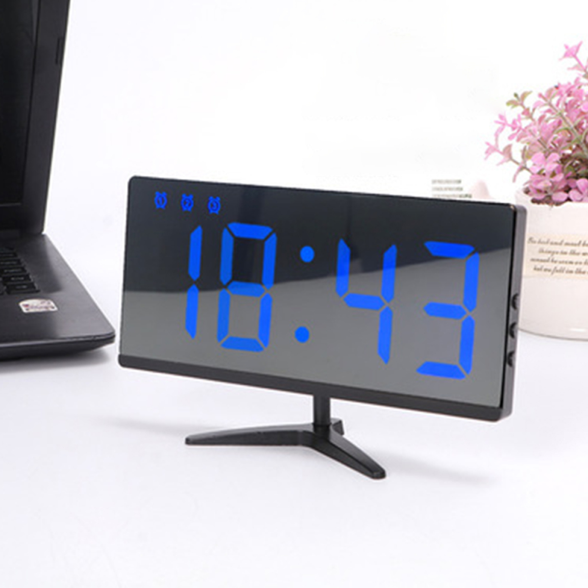 Digitaluhr mit der Anzeige 10:43, mit schwarzem Sockel. Laptop und Pflanze im Hintergrund, weiße Oberfläche.