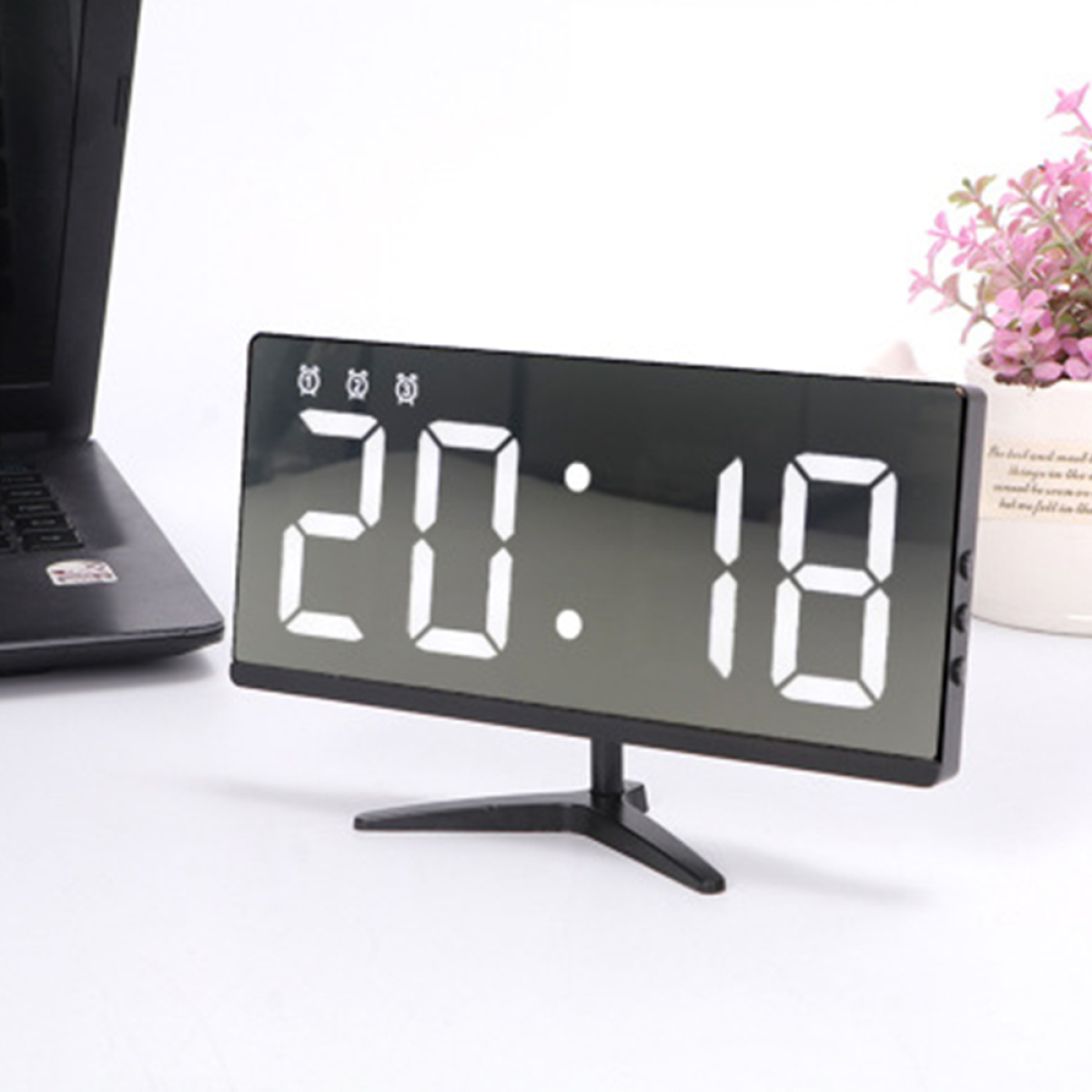 Digitaluhr mit der Anzeige 20:18, auf einem Ständer. Eine Topfpflanze ist in der Nähe.