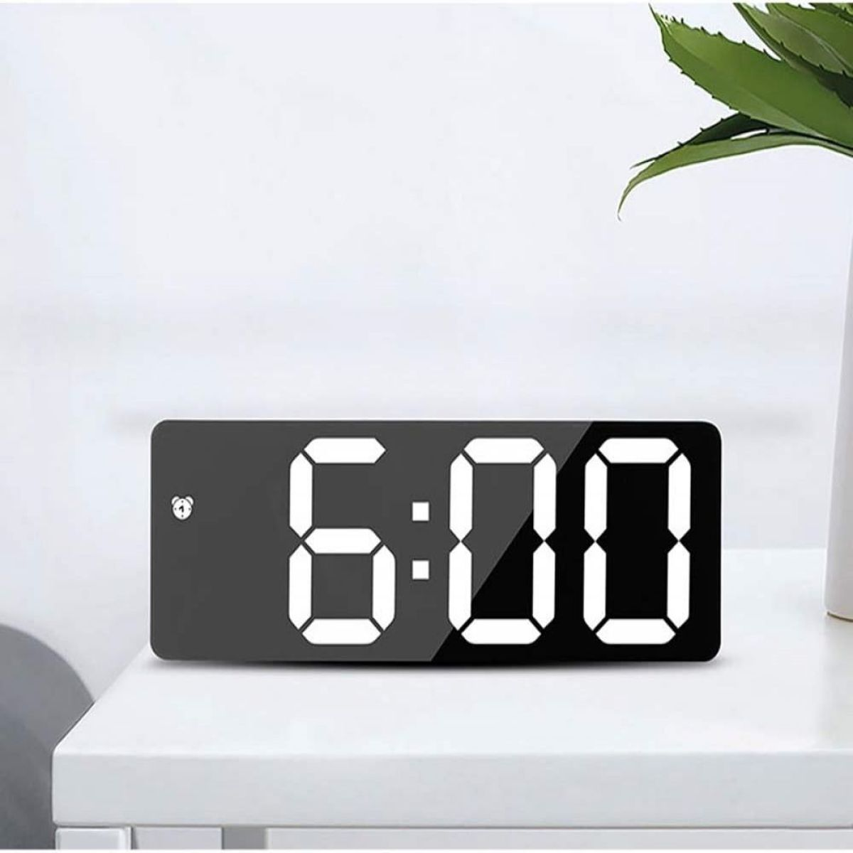 Digitaluhr zeigt 6:00 Uhr auf einem weißen Tisch an. Eine grüne Pflanze steht rechts.