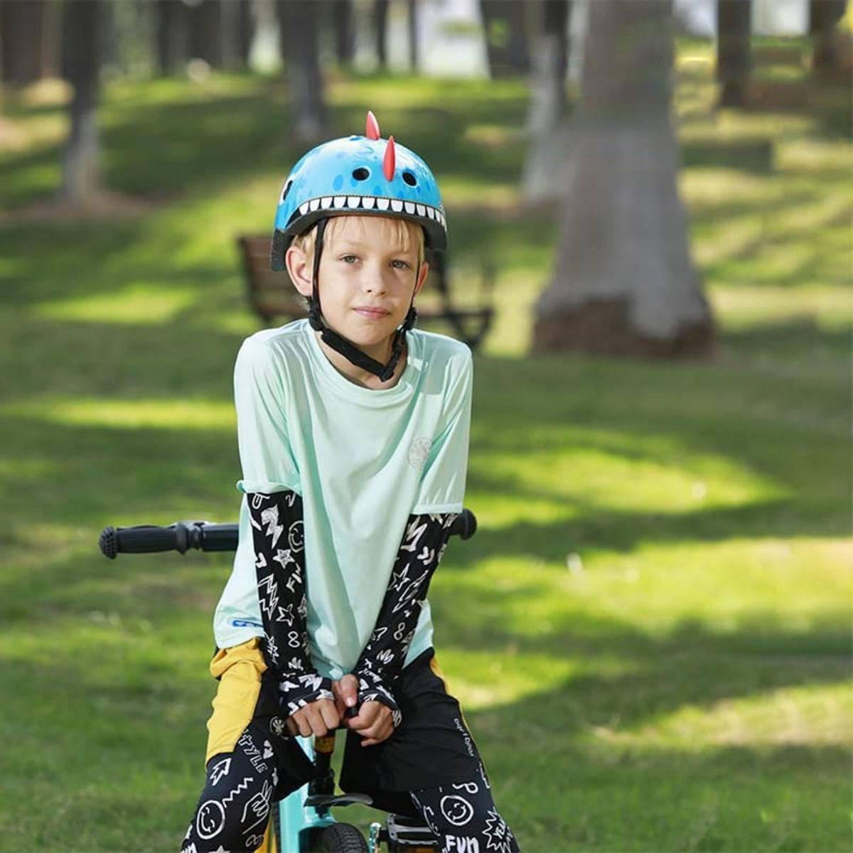 Ein kleiner Junge auf einem Fahrrad, mit blauem Dinosaurierhelm, Ärmeln, in einem Park.