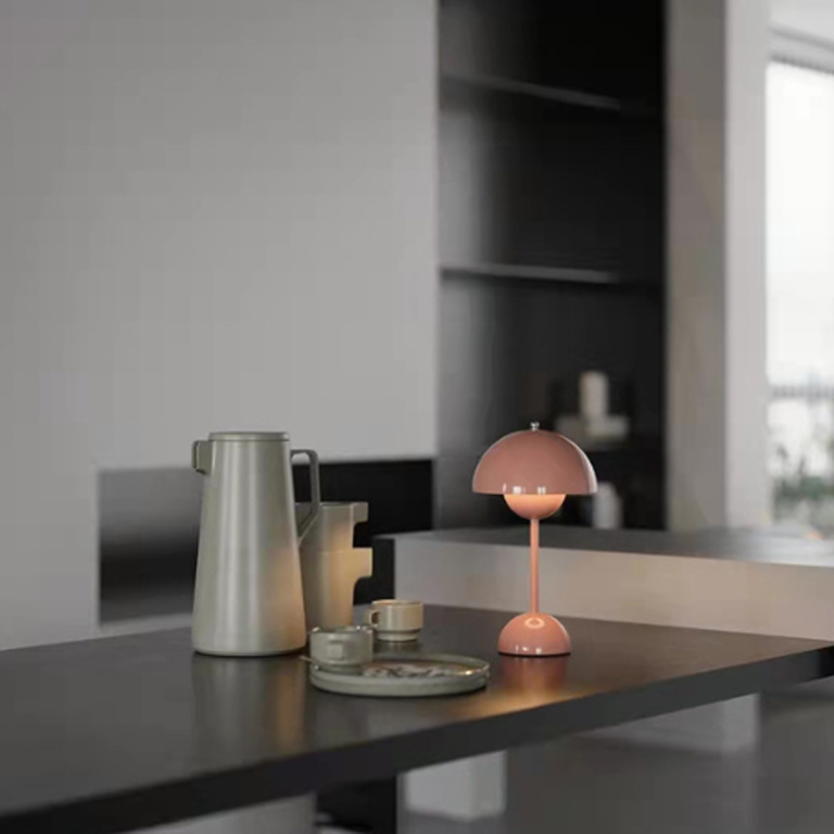 Ein Tisch mit rosa Lampe, weißer Karaffe, Tassen und Teller. Heller, neutraler Hintergrund.