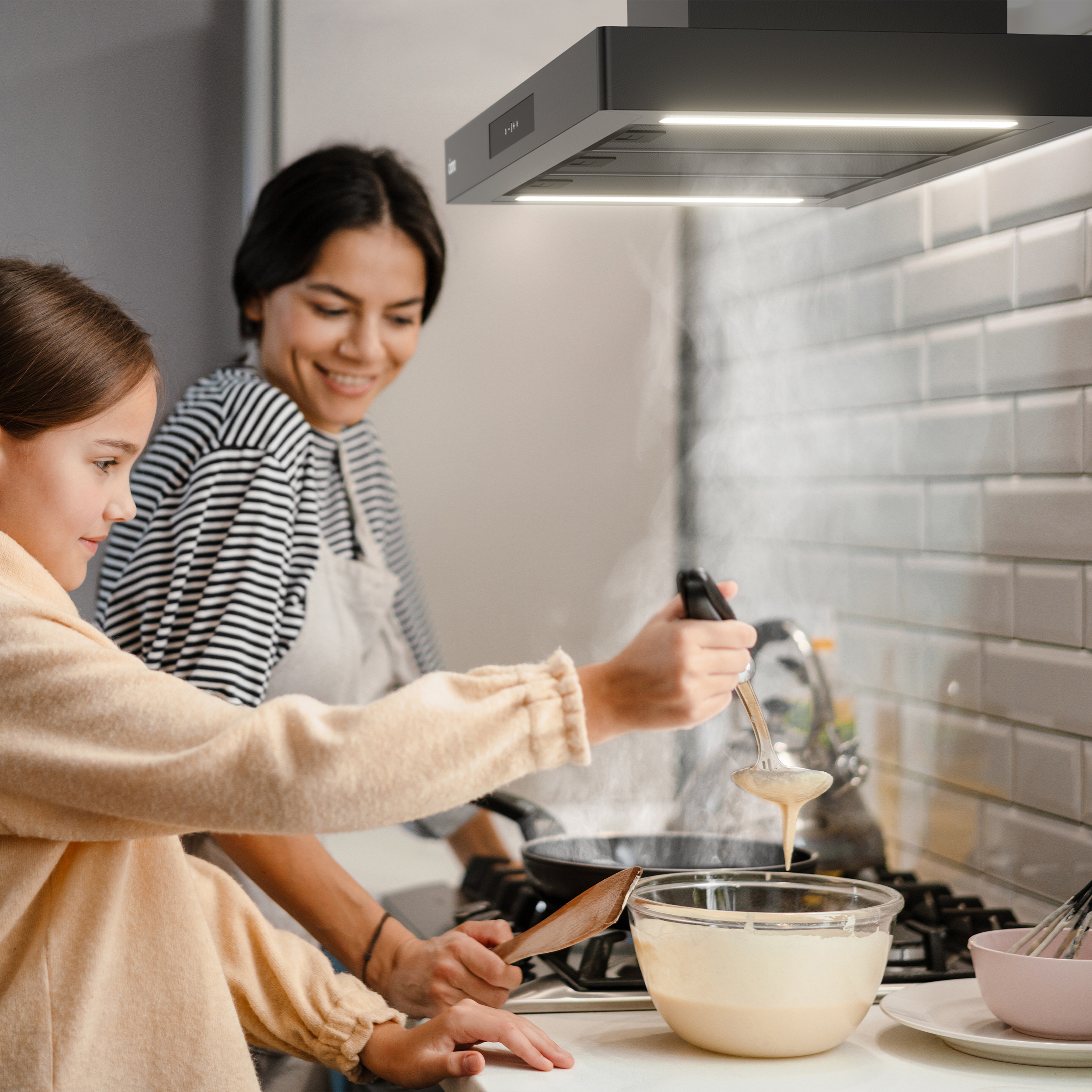 Una donna e una bambina stanno cucinando pancake. Vapore sale sopra la padella. Ambiente cucina.