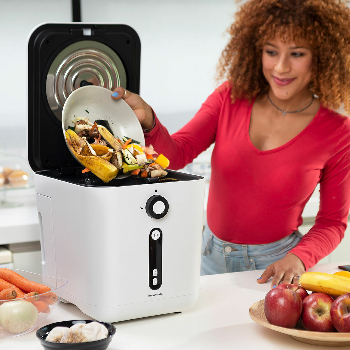 Mujer vertiendo verduras de un cuenco a un compostador de alimentos blanco, con manzanas y cebollas cerca.