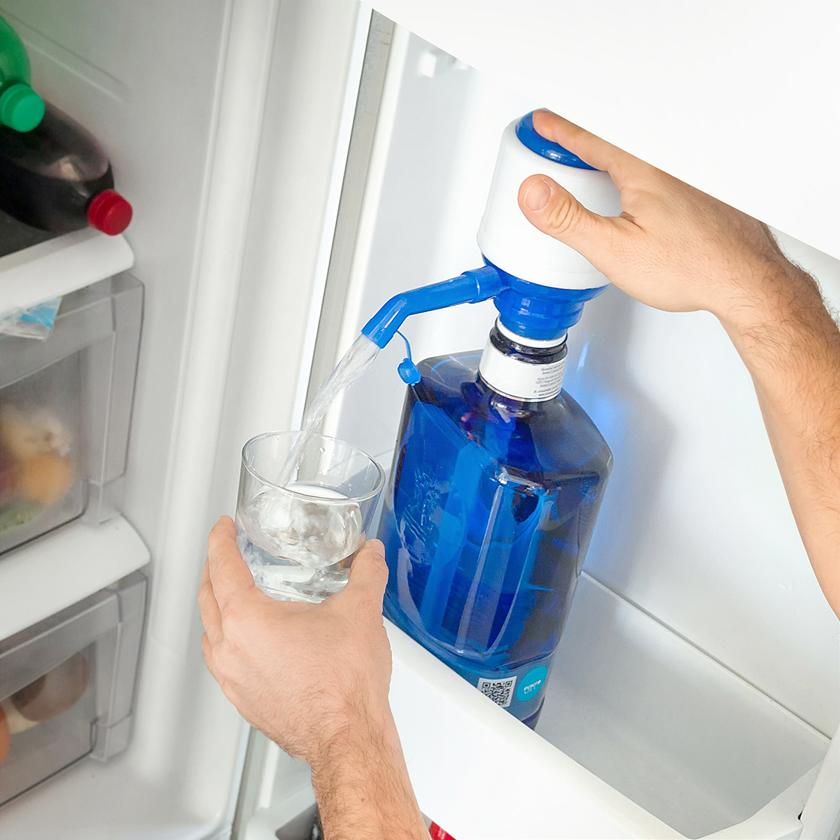 Eine Person benutzt eine Pumpe, um ein Glas mit Wasser aus einer blauen Wasserflasche in einem Kühlschrank zu füllen.