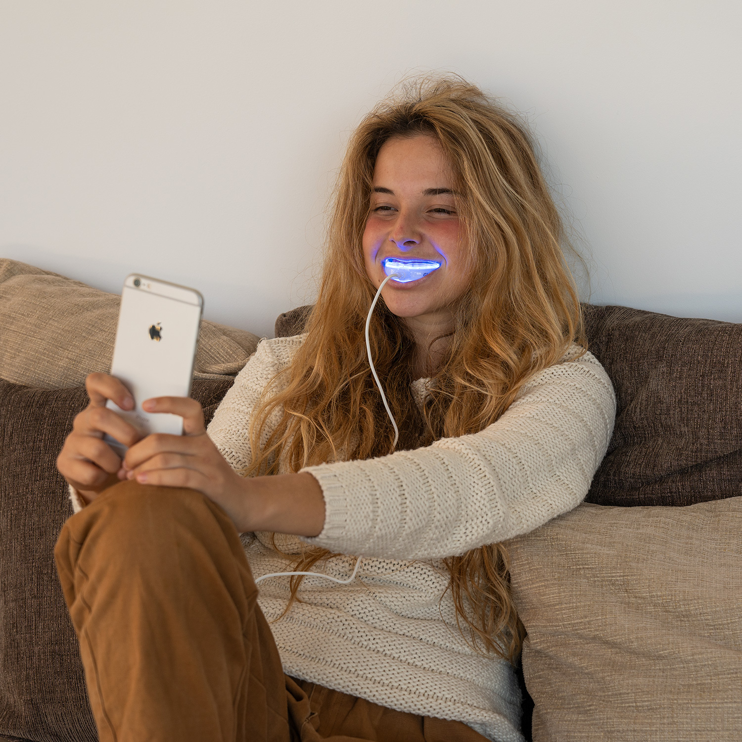 Mujer usando smartphone con dispositivo blanqueador dental de luz azul. Sonríe en un sofá.