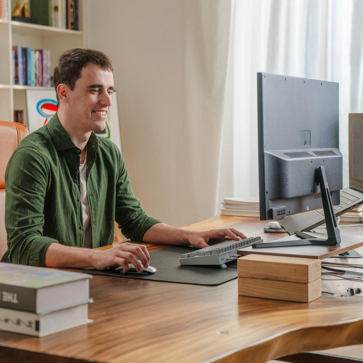 Mann lächelt am Computer. Schreibtisch mit Büchern, Maus, Tastatur und Monitor.