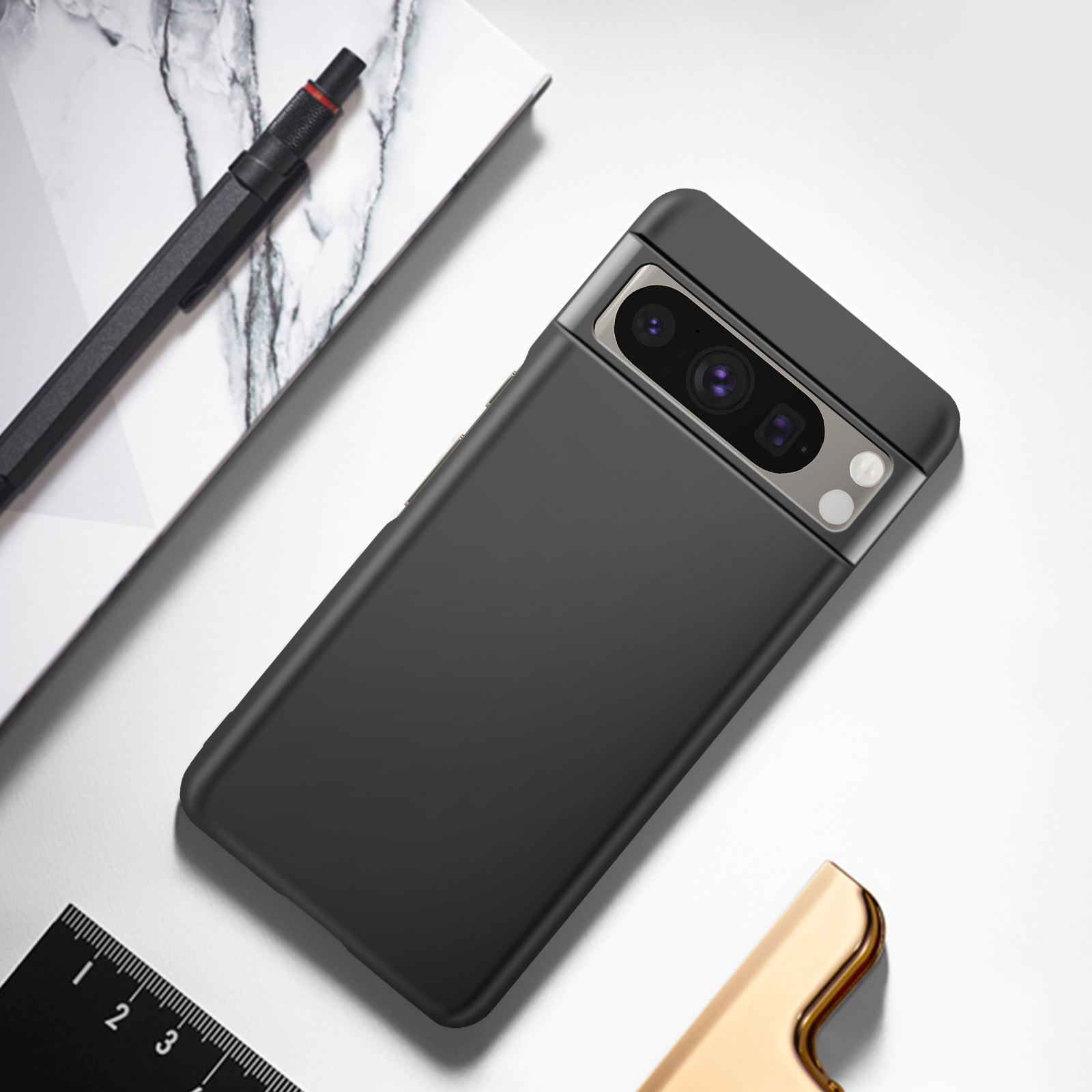 Telefono nero con fotocamera, custodia, righello e un accendino dorato su una superficie bianca.