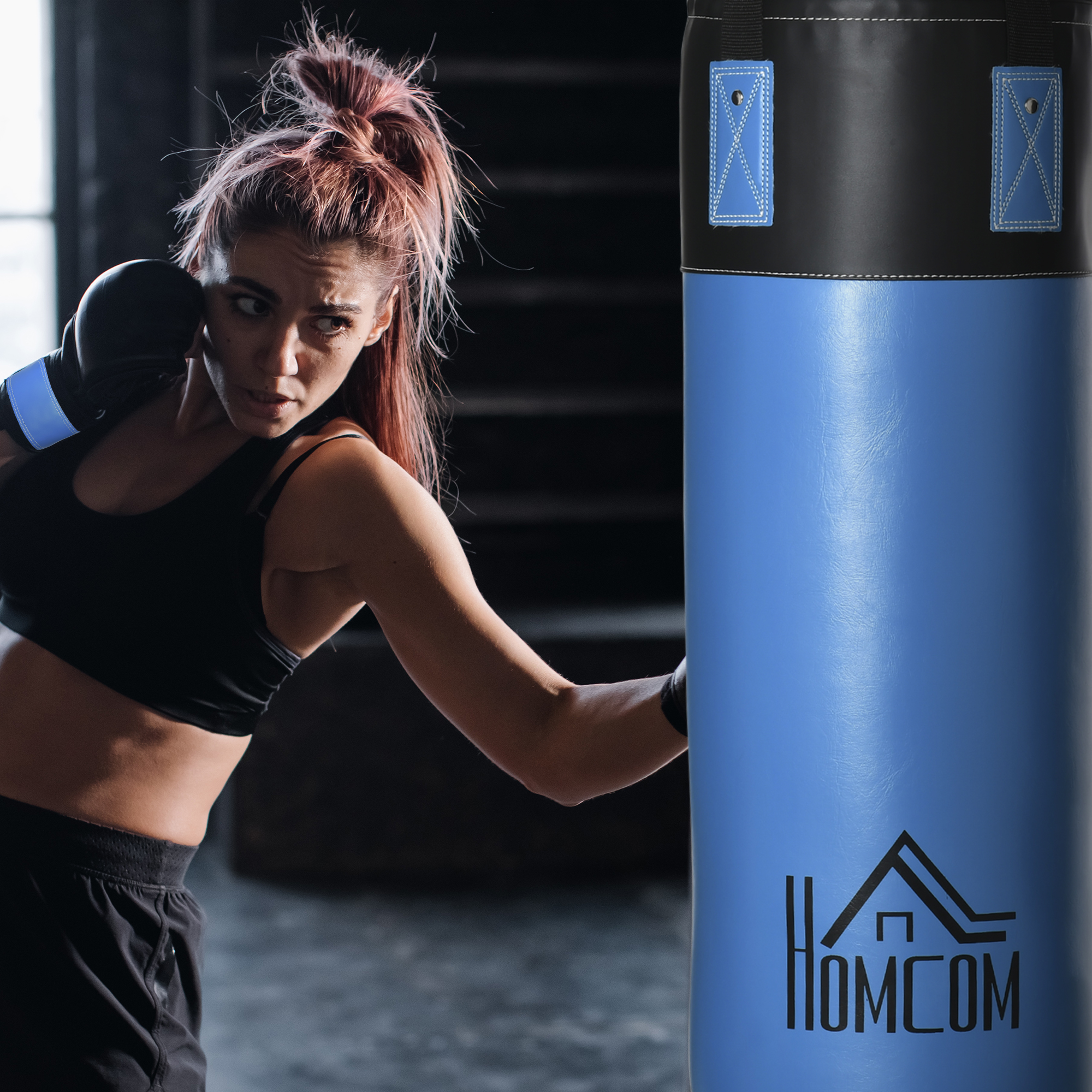 Mujer boxea un saco de boxeo azul. Usa guantes negros y un sujetador deportivo en una habitación oscura.
