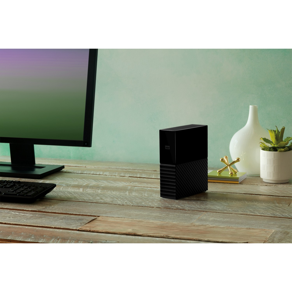 Czarny zewnętrzny dysk twardy na drewnianym biurku obok monitora, wazonu i rośliny.