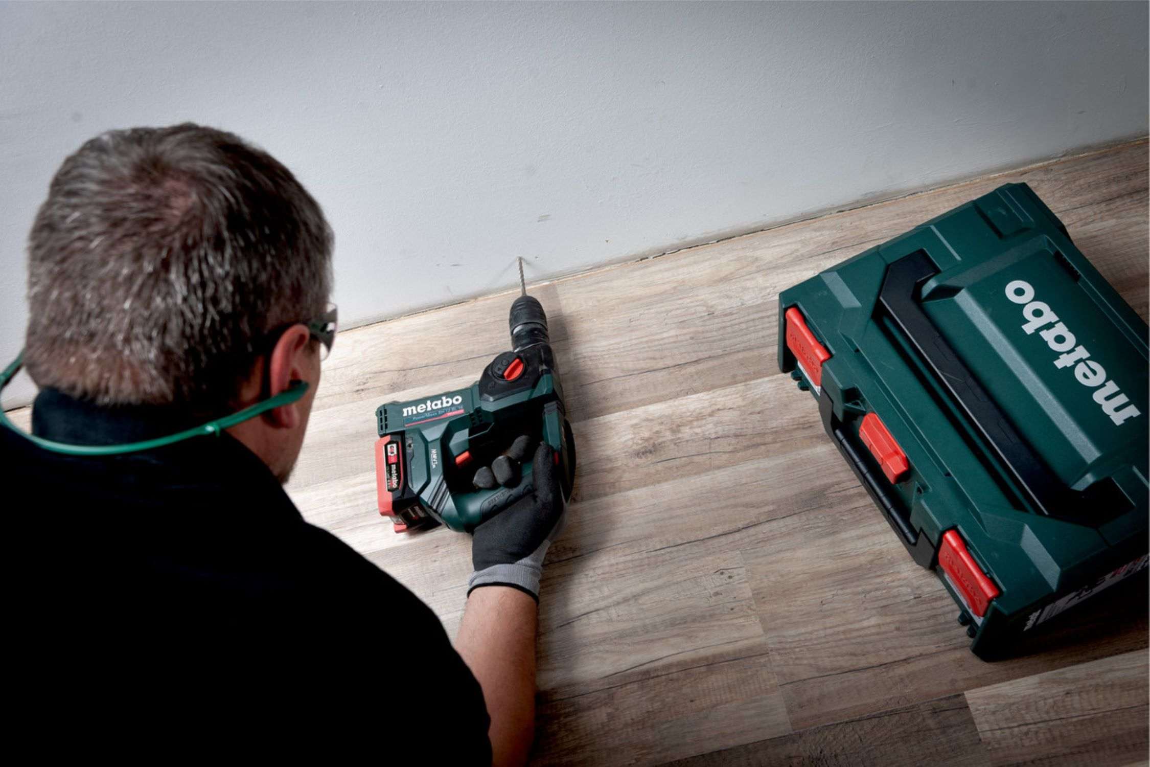 Person bohrt mit einer Metabo-Bohrmaschine in eine Wand, neben einem grünen Werkzeugkoffer.